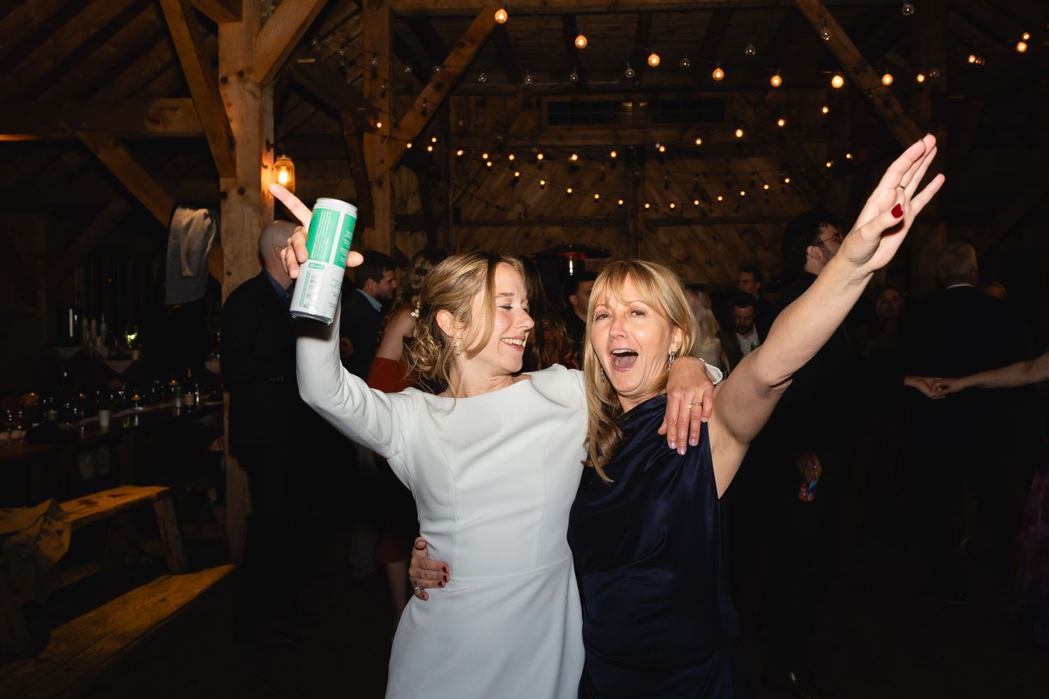 Two joyful party attendees dancing together at a rustic barn venue with string lights overhead during a celebration.