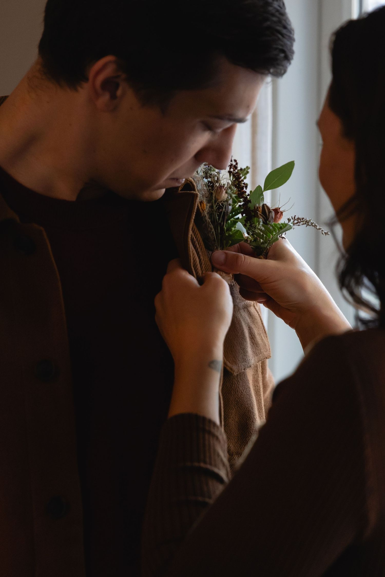 Intimate moment between couple as person pins a small floral boutonniere to their partner's brown sweater.