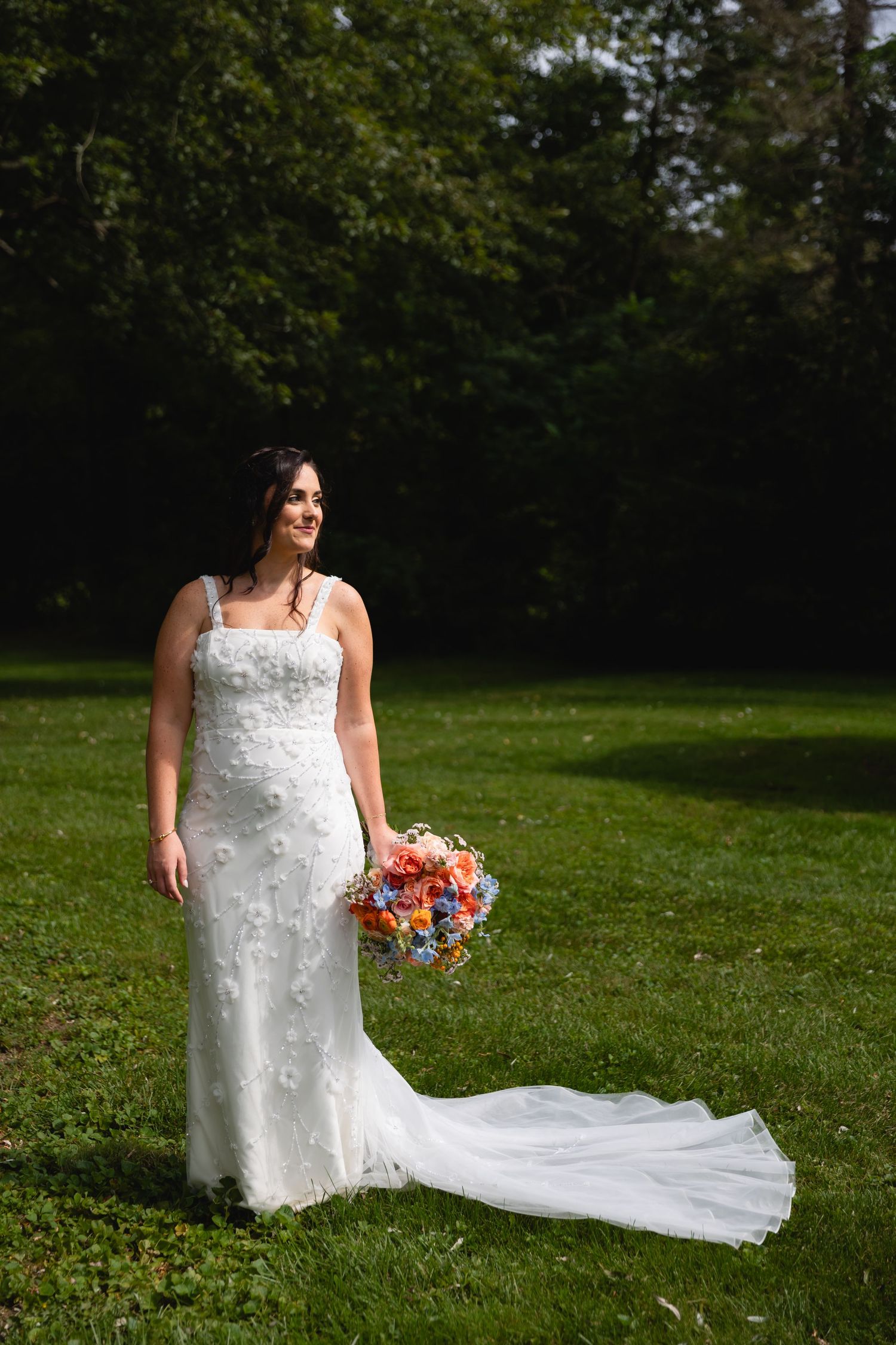 A bride in a white lace gown with train holds a vibrant coral and pink bouquet while standing on green grass.