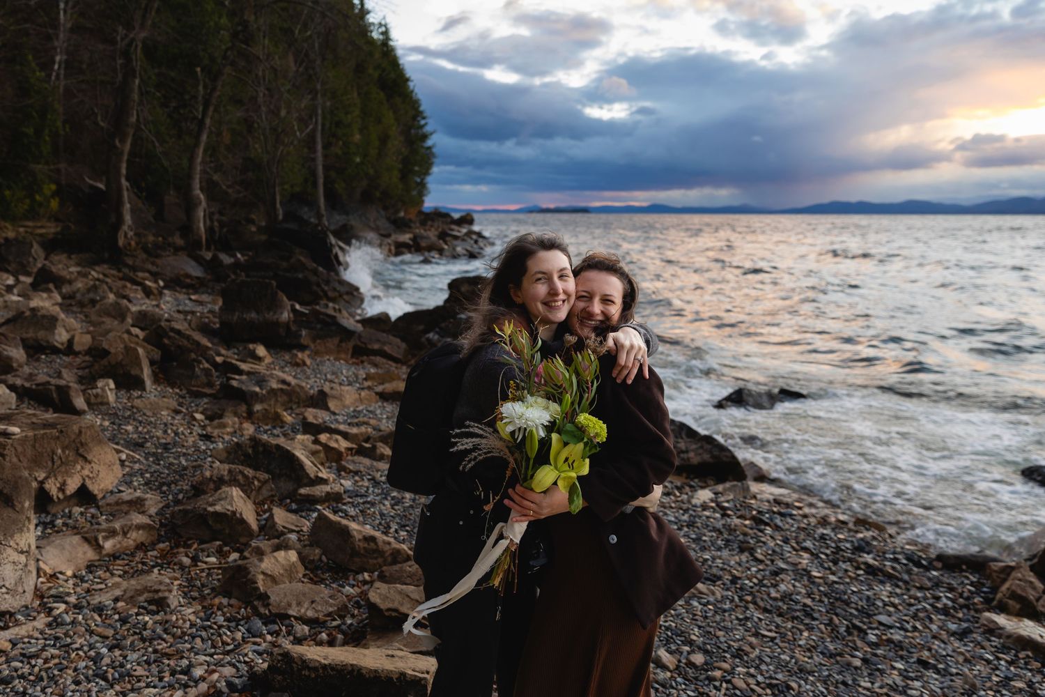 Two people in dark clothing share an intimate moment on a rocky beach at sunset with stormy clouds overhead.