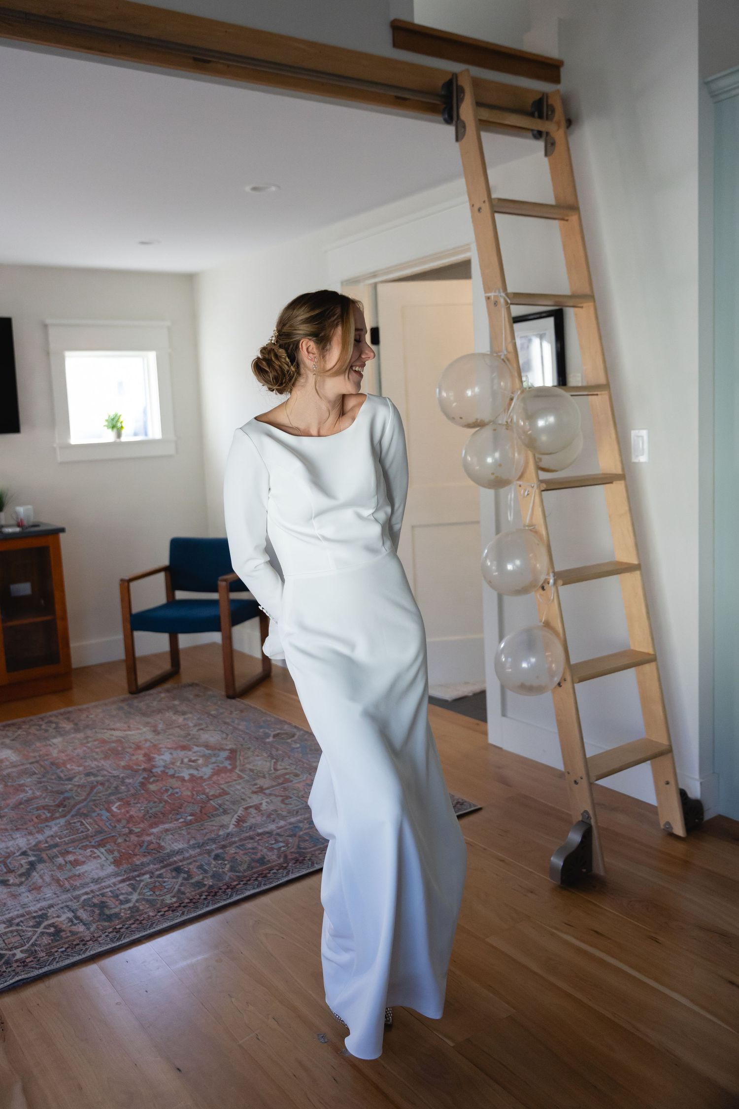 A figure in an elegant white flowing dress stands in a modern minimalist room with wooden ladder and glass orb decorations.