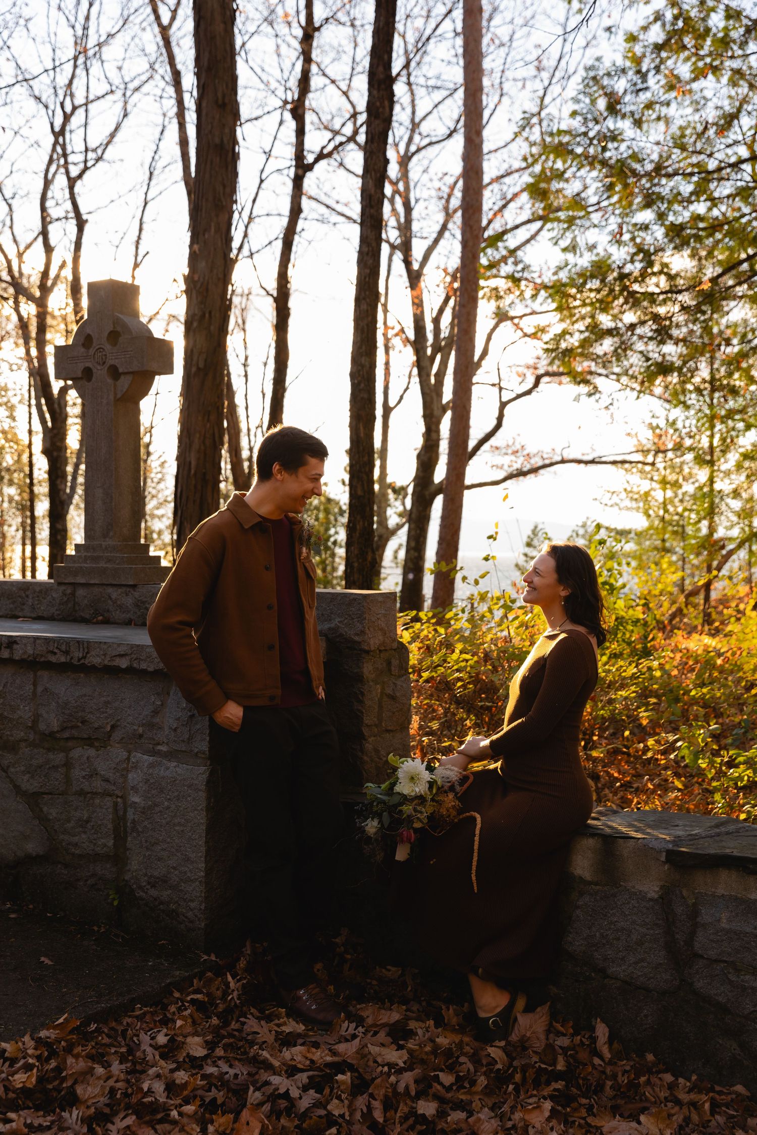 Two people in dark clothing converse near a stone wall with a Celtic cross and autumn trees in warm sunset light.