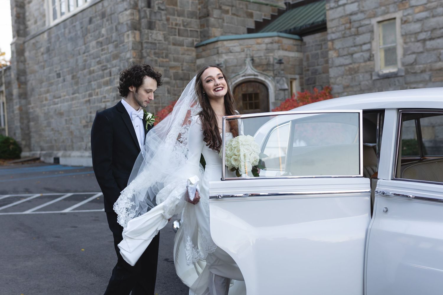 A bride in a flowing white gown exits a vintage white car outside a stone building on their wedding day.