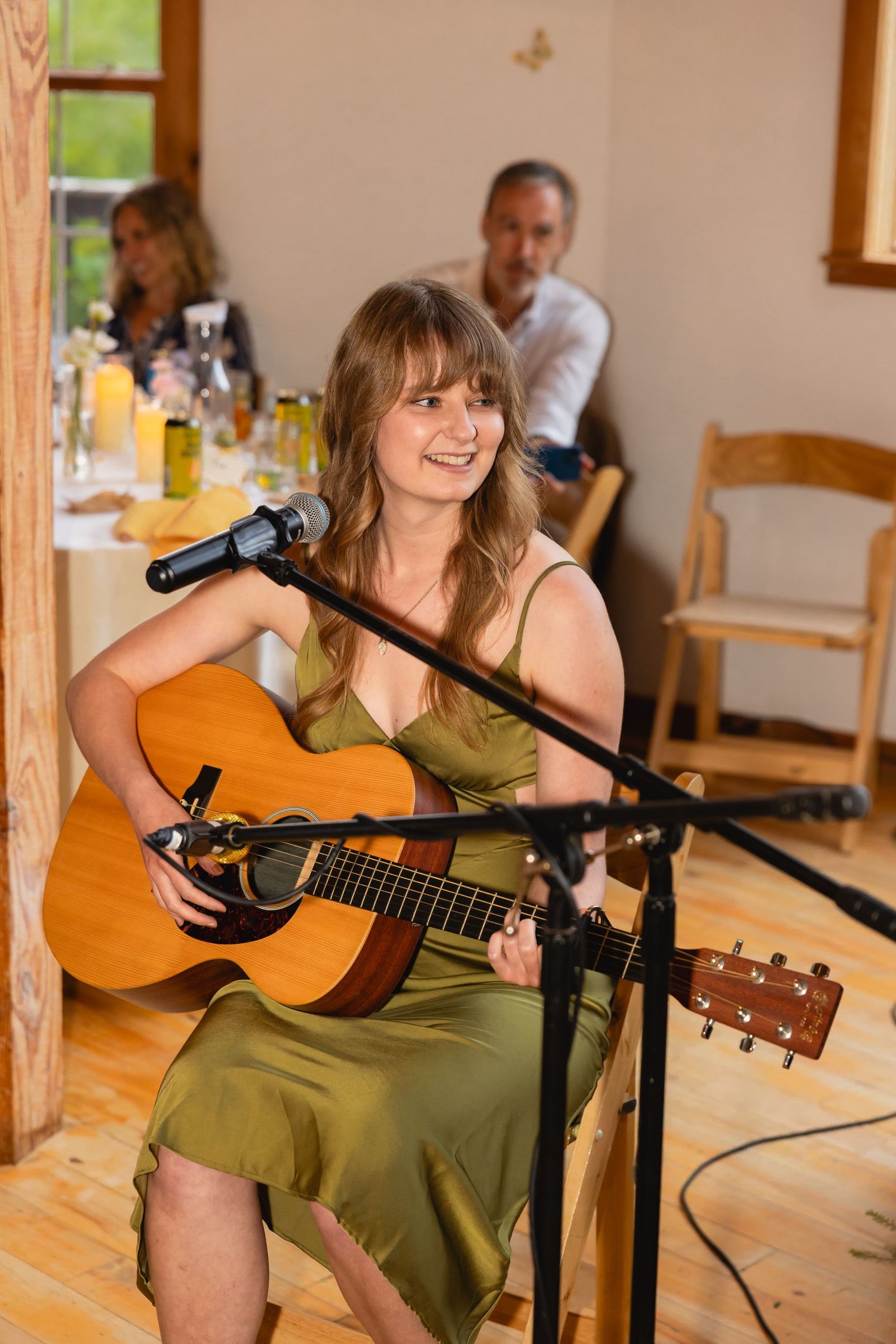 Musician in olive green dress performs with acoustic guitar at indoor venue.