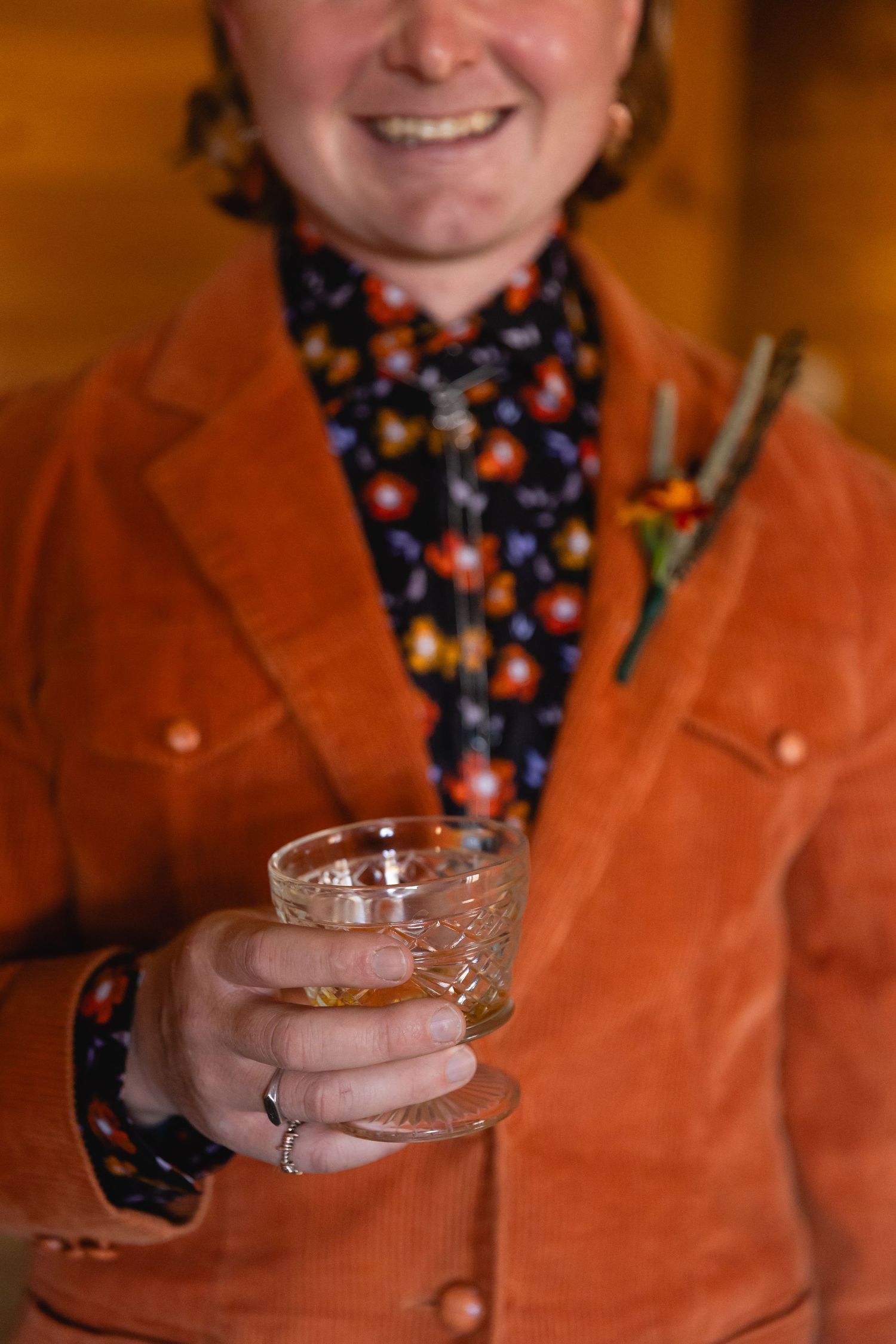 Person wearing an orange corduroy jacket and floral print scarf holds a coupe cocktail glass.