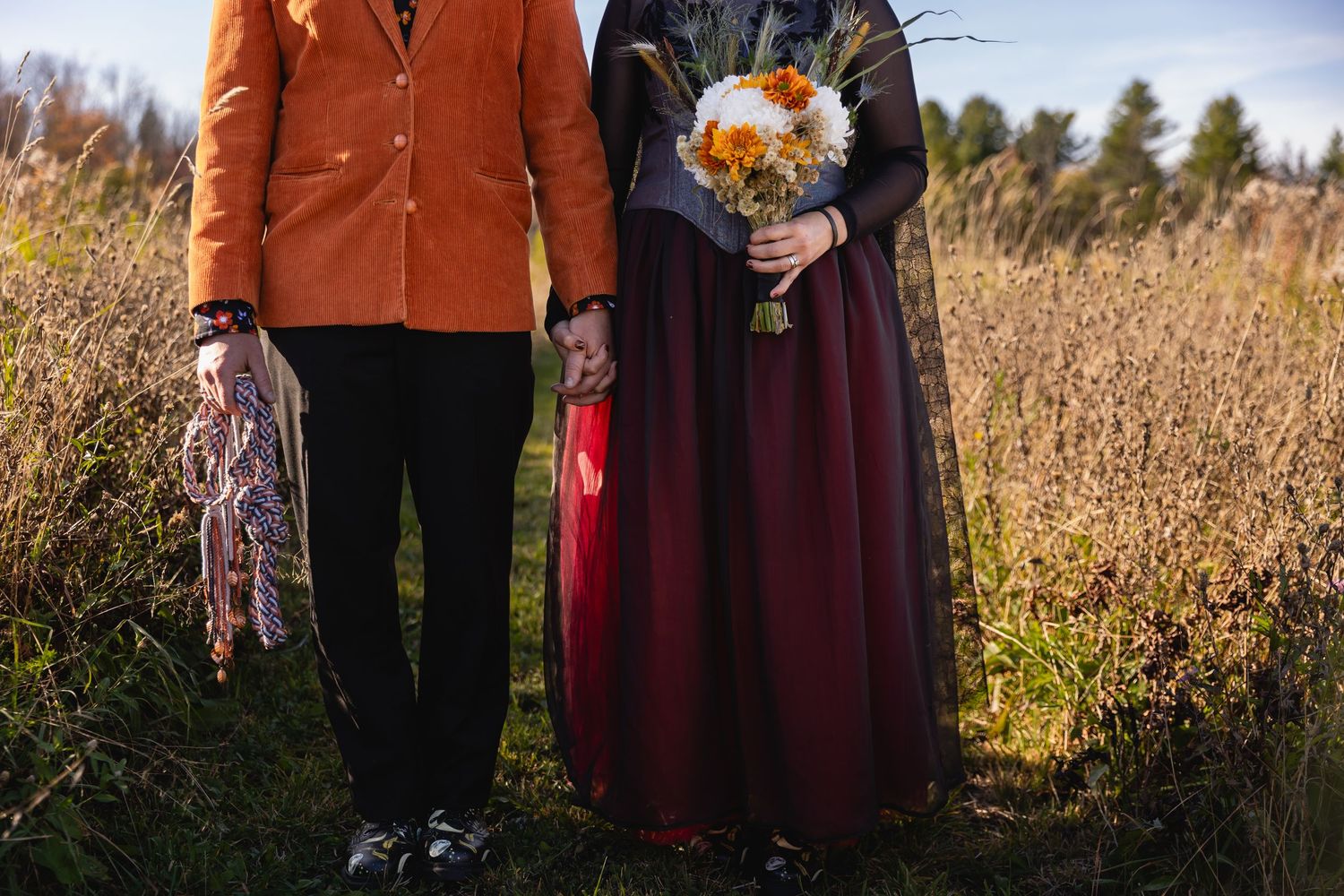 A couple stands in a fall field wearing orange and burgundy clothing while holding a rustic autumn bouquet.