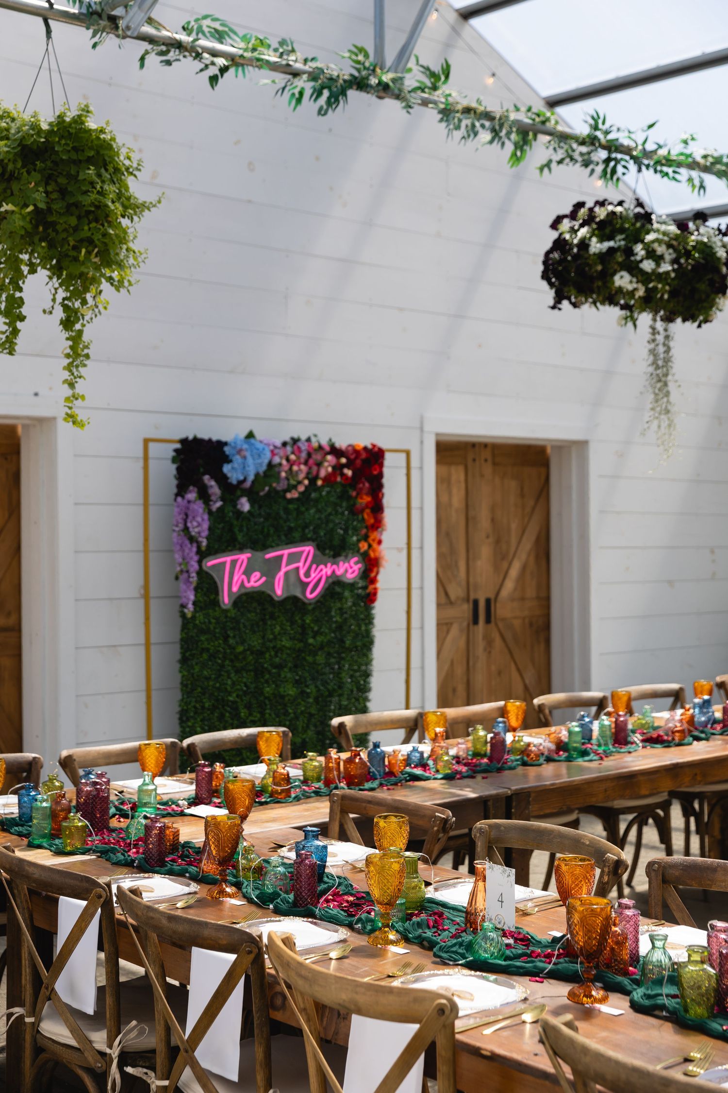 A long rustic wooden table set for a party with hanging plants and a neon pink sign against a white wall.