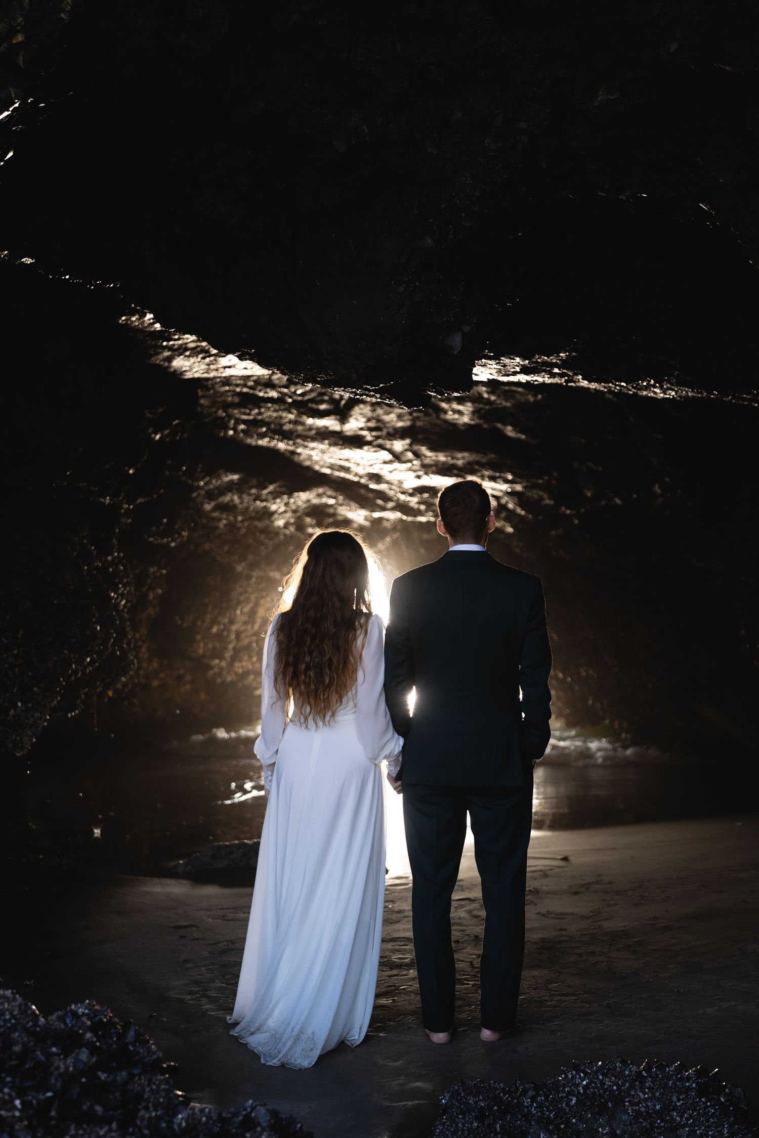 A couple stands silhouetted in a dark cave with dramatic backlighting creating a moody atmosphere.
