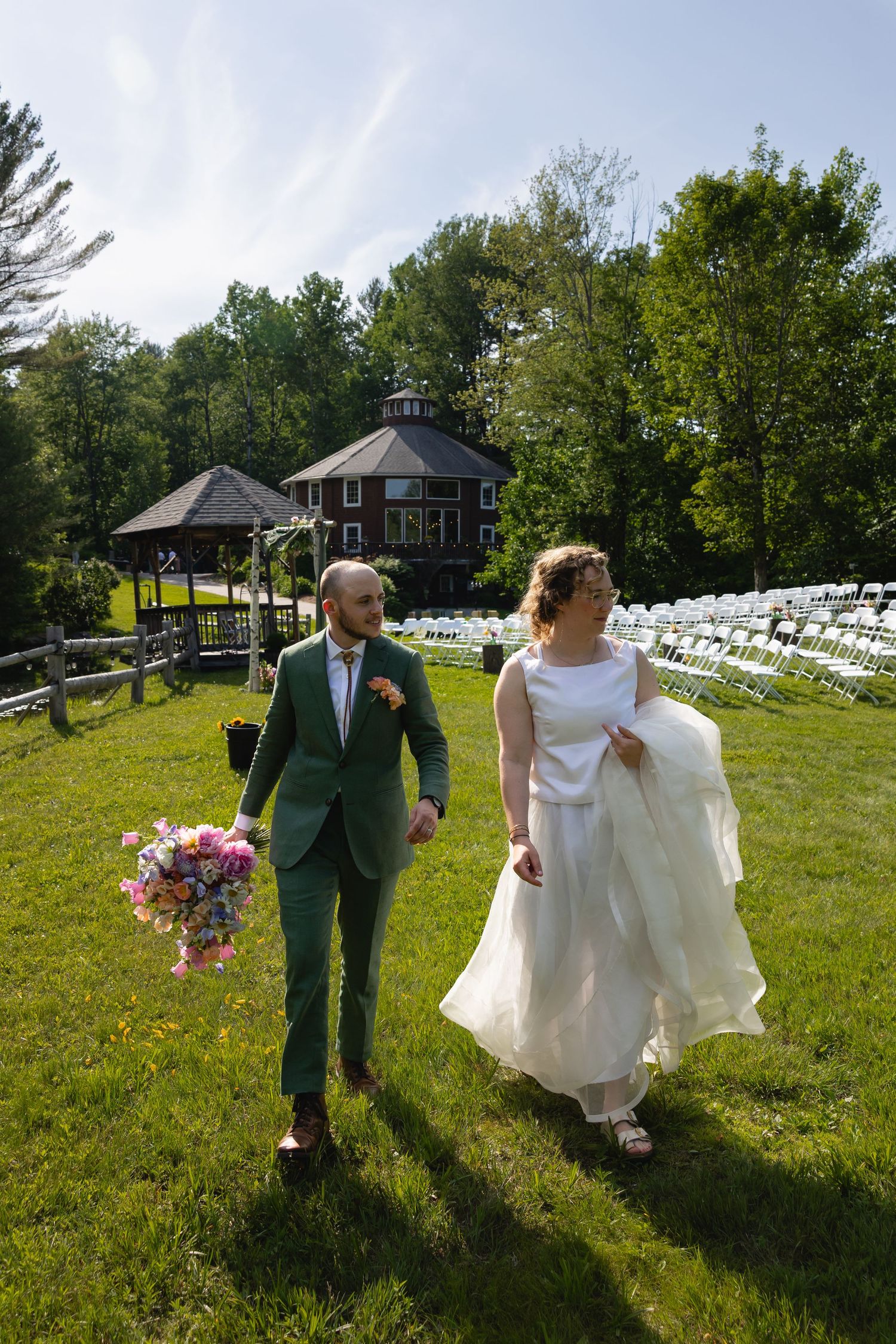 A couple walks together across a sunny lawn with white chairs and a rustic building in the background.