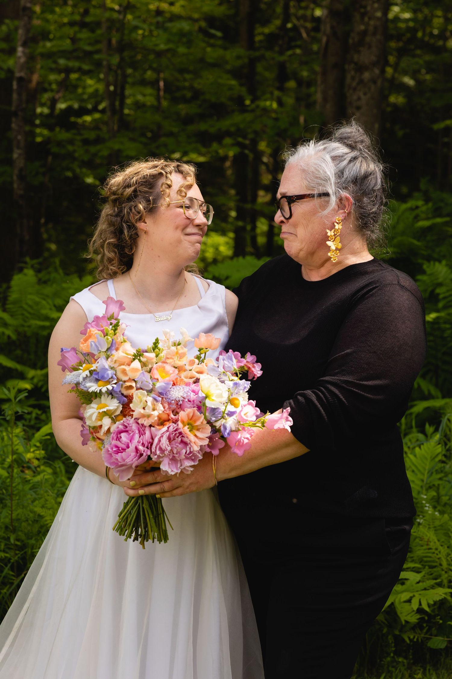 A couple shares a joyful moment in a lush forest setting while holding a vibrant pink and peach floral bouquet.