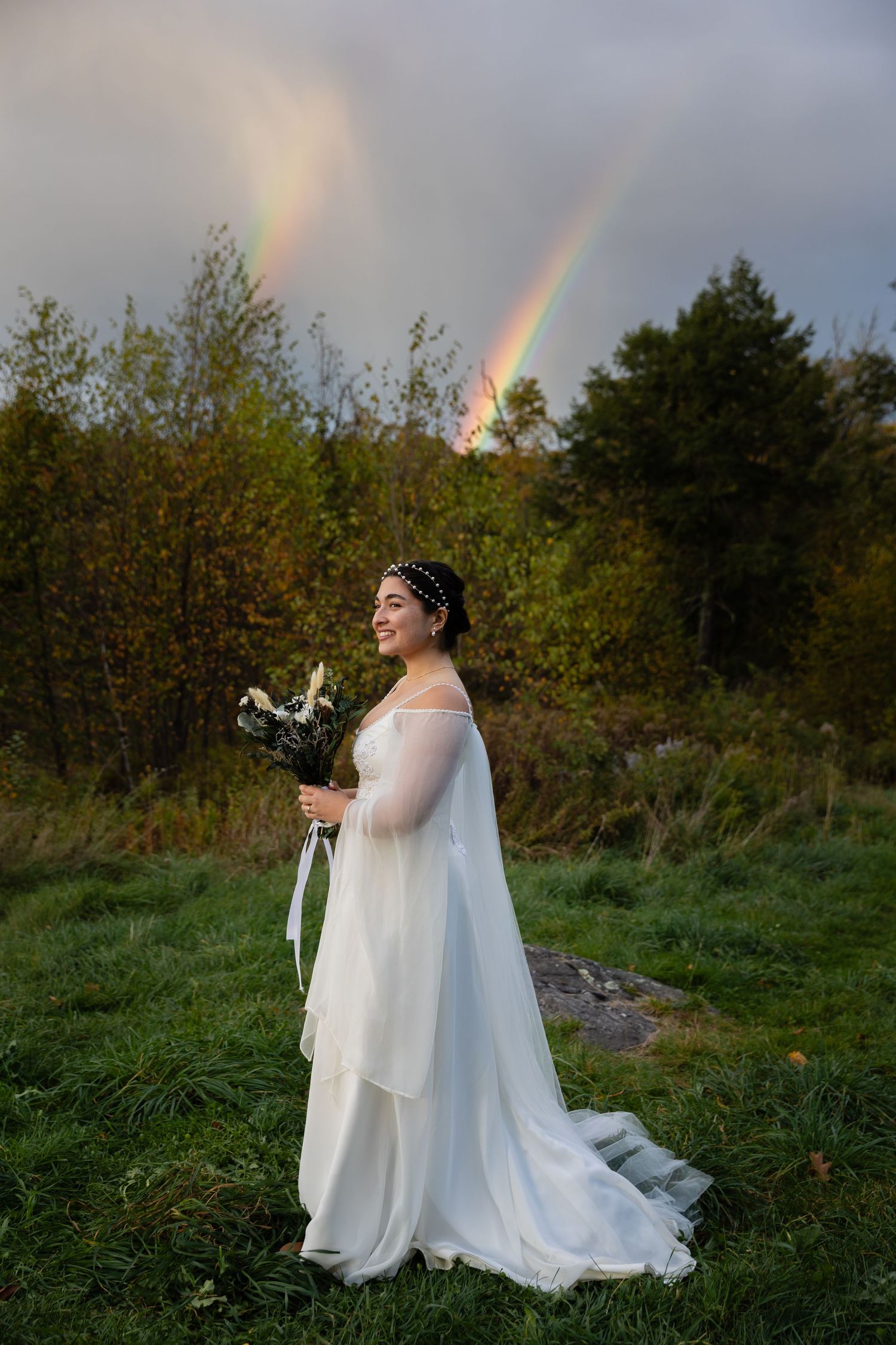 A bride in a flowing white dress stands holding a bouquet with a rainbow in the background.