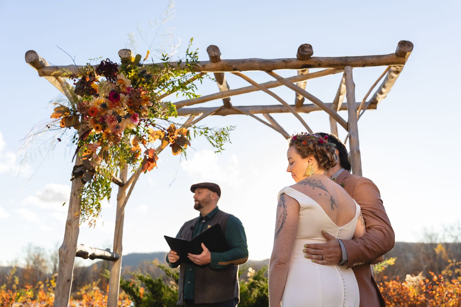 An outdoor wedding ceremony under a wooden arch decorated with autumn flowers and foliage.
