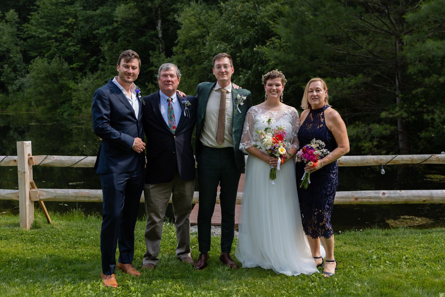 Wedding group photo with bridal party standing by wooden fence in garden.