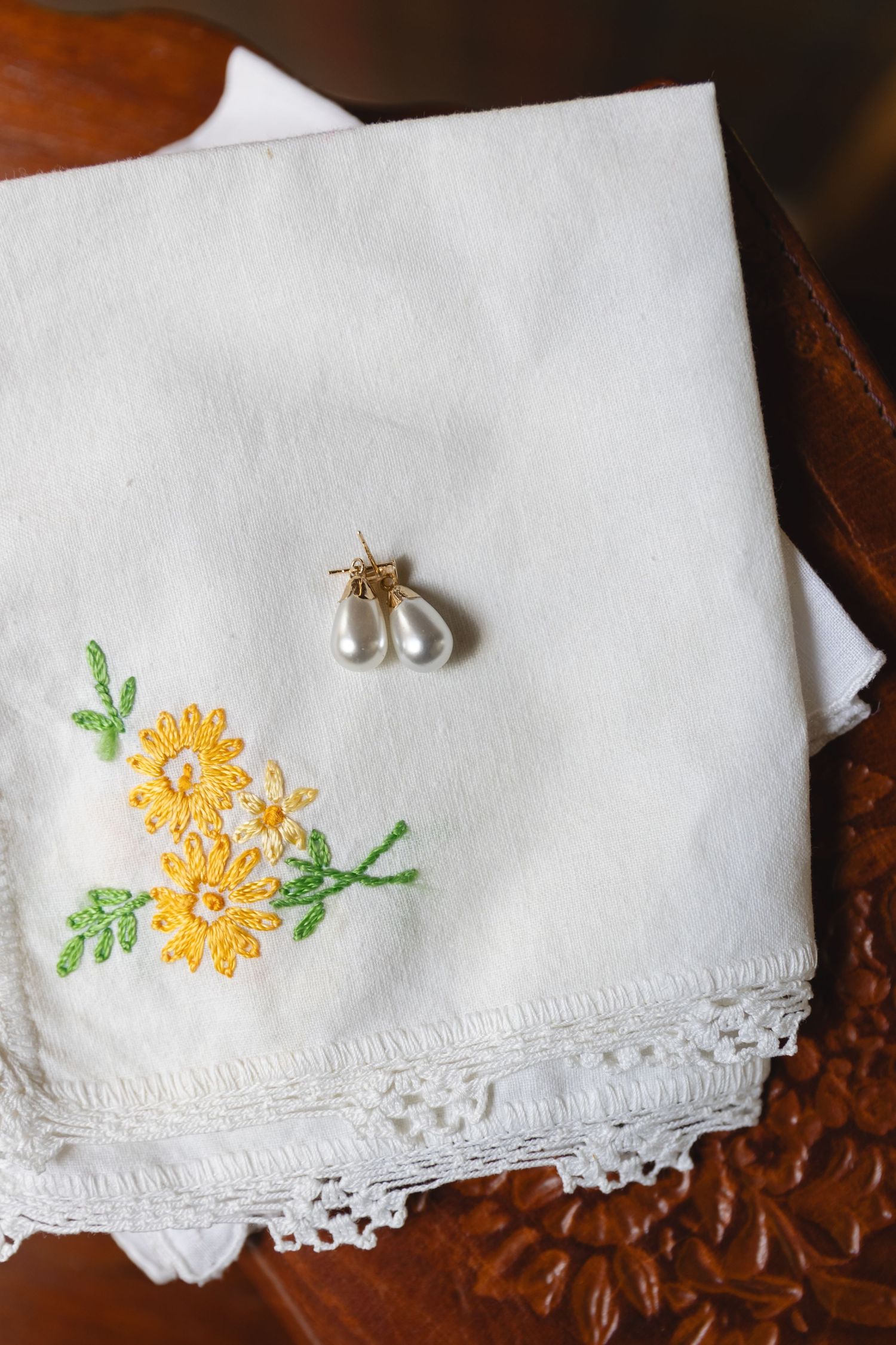 Delicate embroidered handkerchief with yellow flowers and pearl earrings against dark wood.