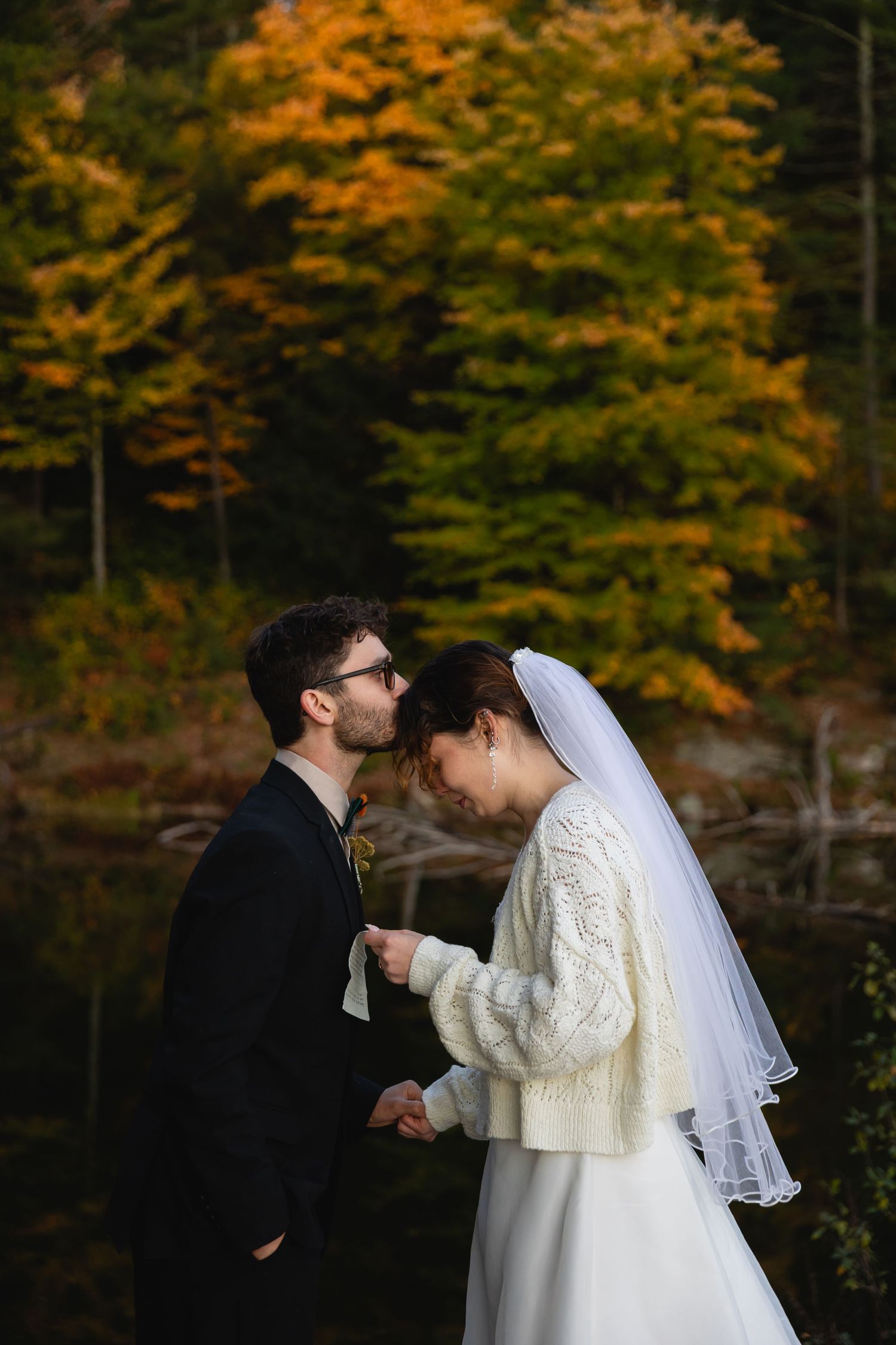 Groom kisses bride's forehead near autumn foliage lake, bride wearing cozy white knit sweater.