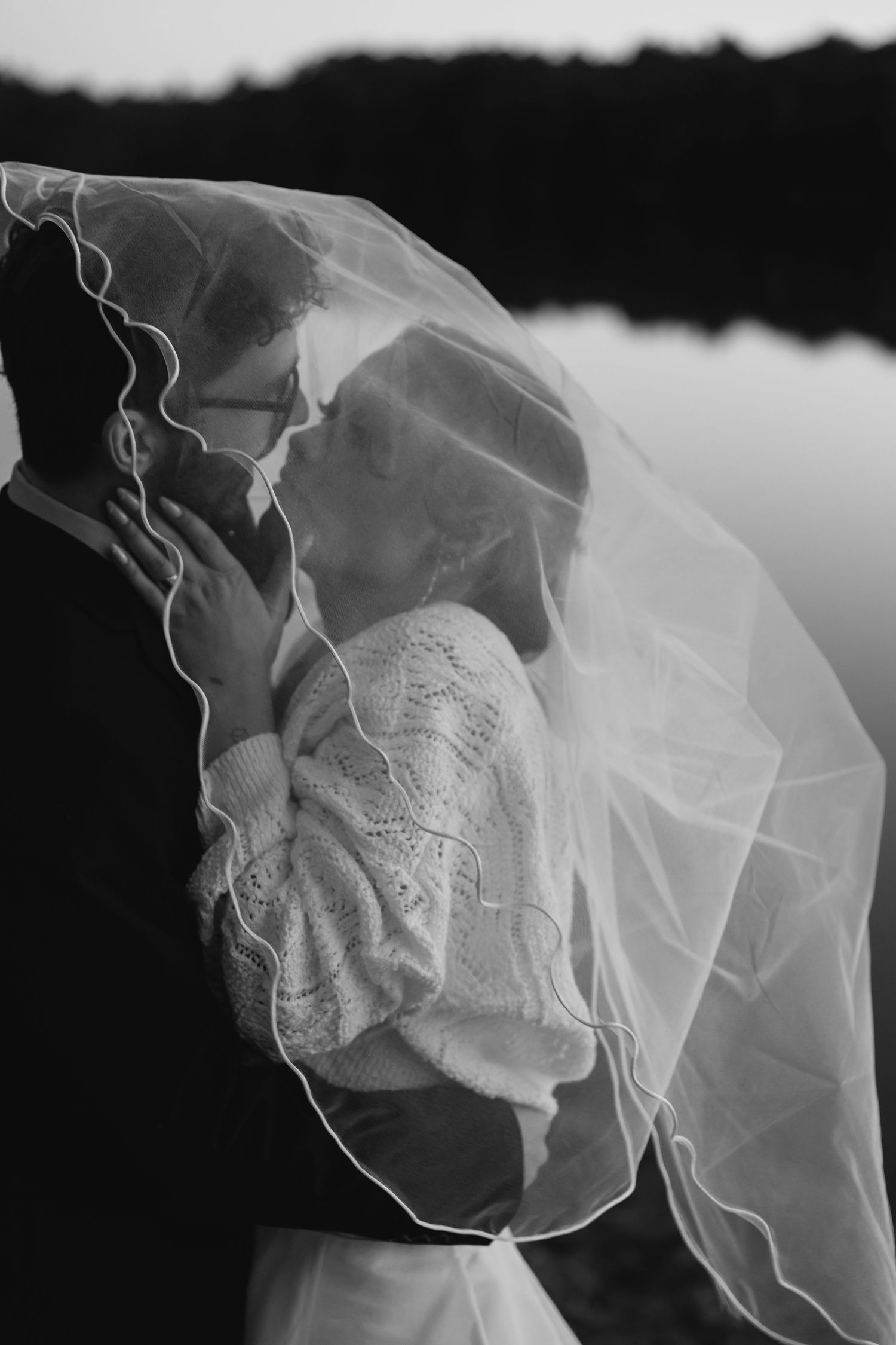 Black and white romantic photo of a bride and groom kissing beneath a flowing veil by a serene lake.