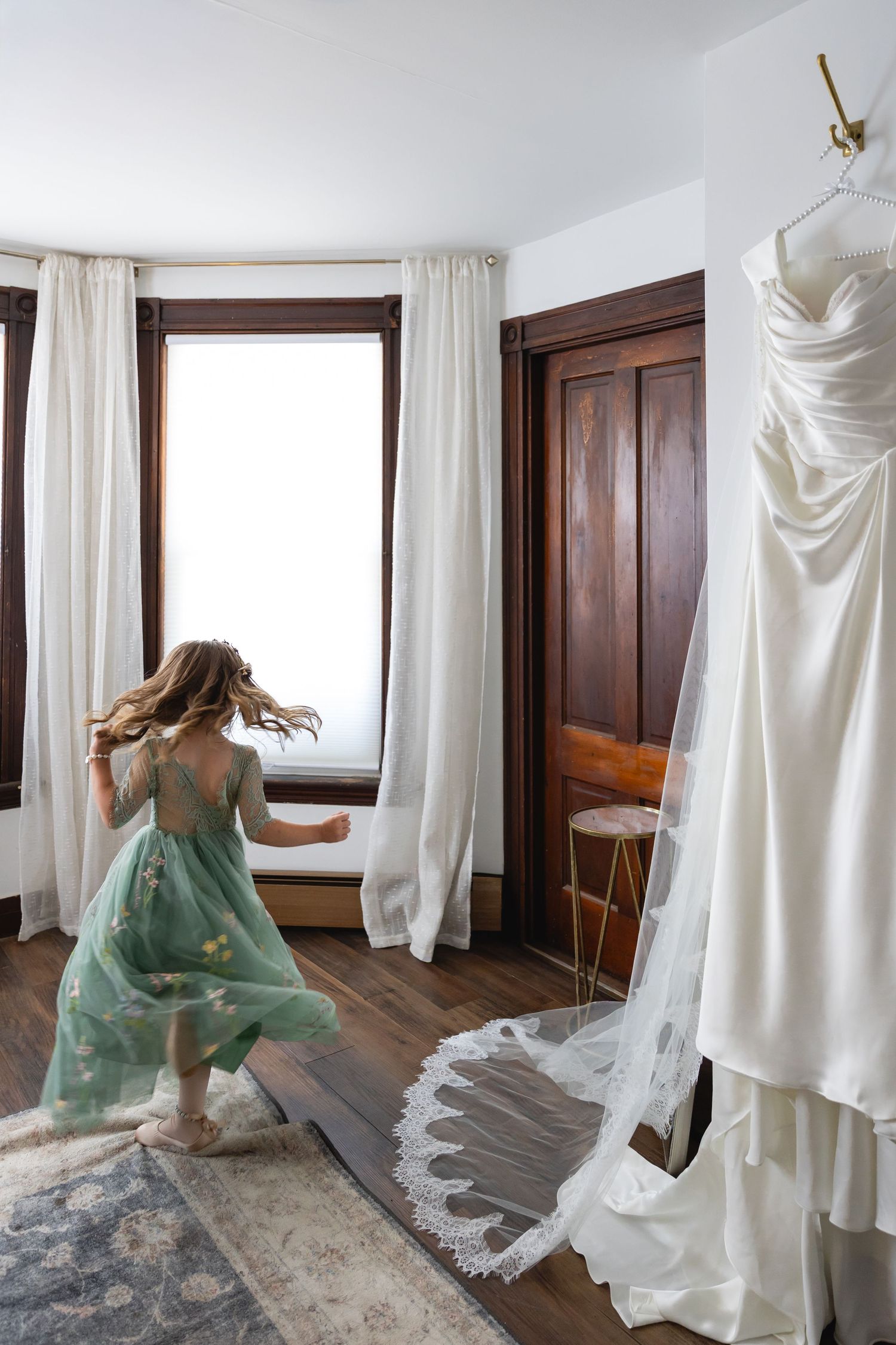 Flower girl in green dress twirling near a hanging wedding gown in a bright bridal suite.