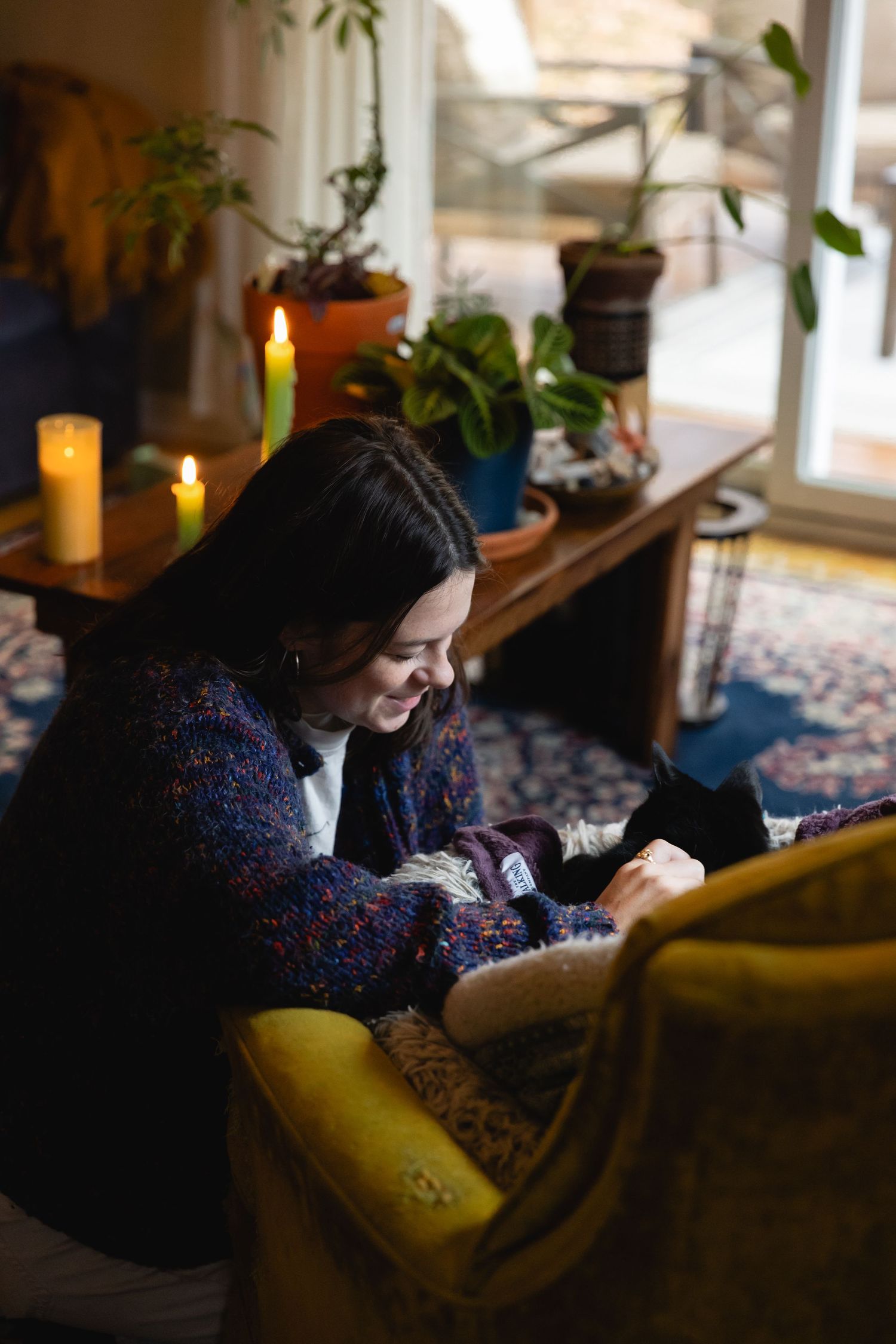 Person relaxes in cozy living room corner with glowing candles and houseplants, reading on a yellow armchair.