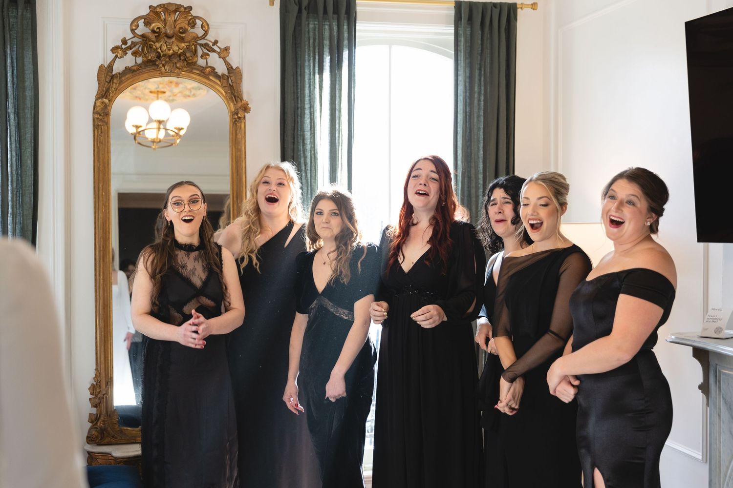 A group in black dresses share a candid laughing moment together in an elegant room with ornate gold mirror.
