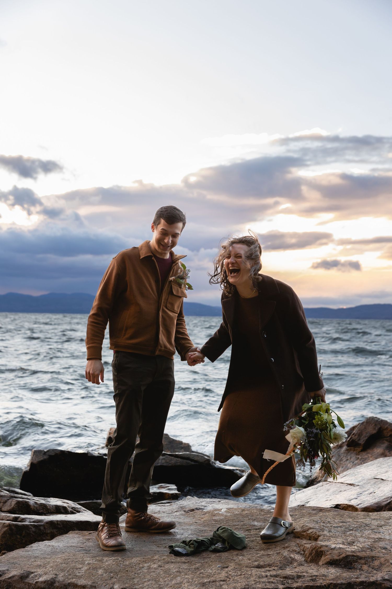 Two people share a joyful moment on rocky coastal cliffs at sunset with ocean waves crashing in the background.