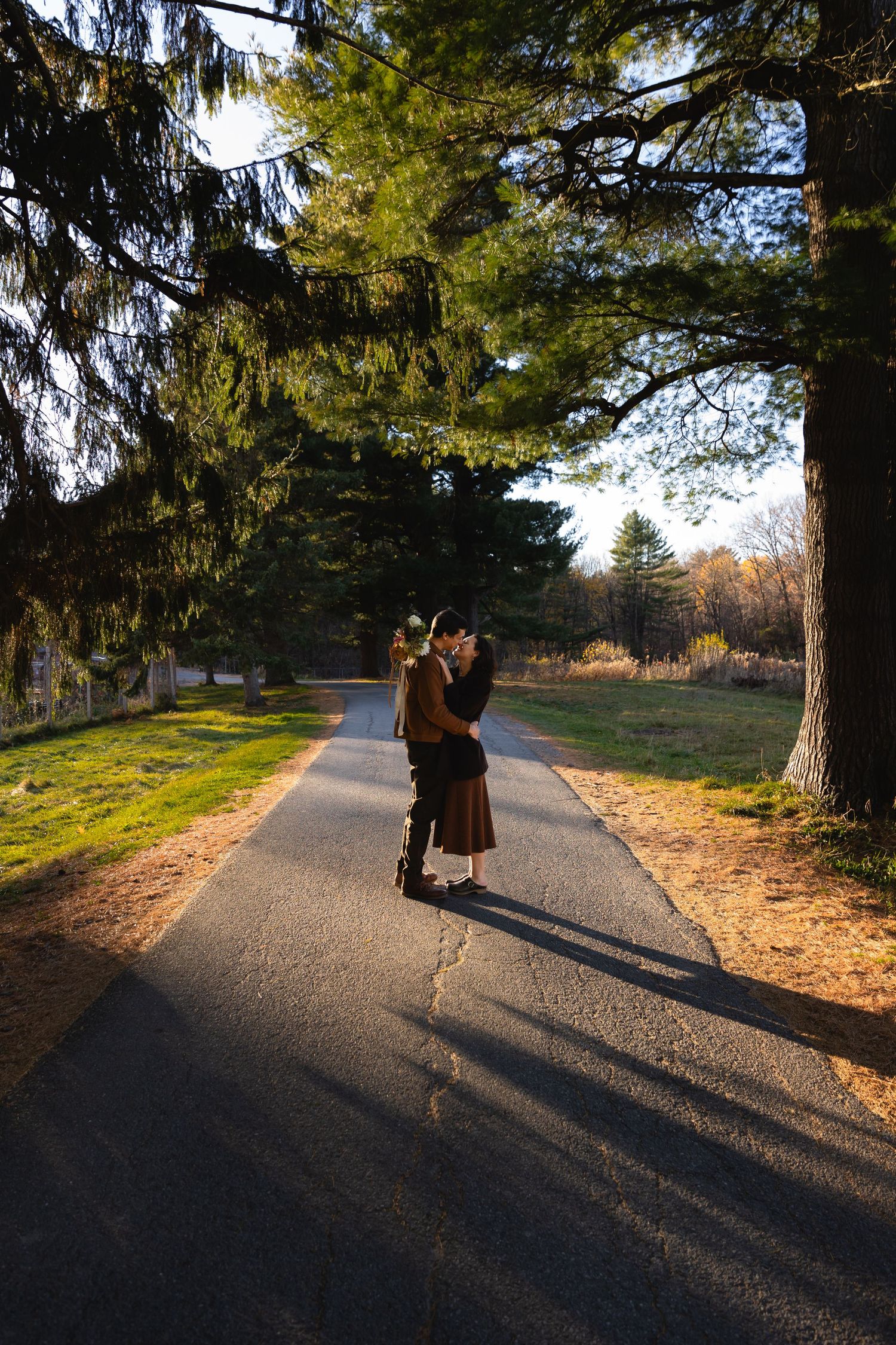 A silhouetted couple stands together on a tree-lined path during golden hour.