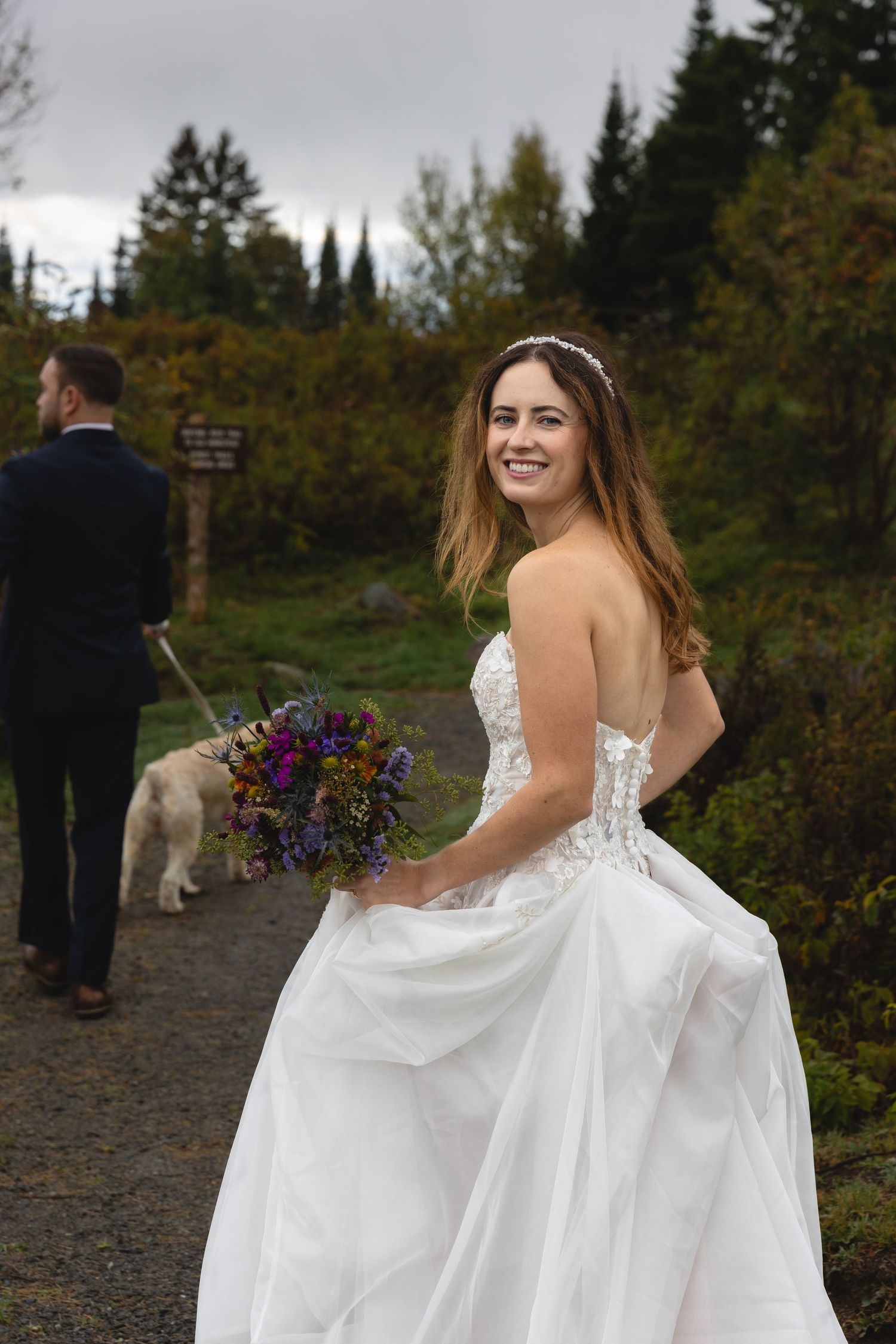 A bride in a white lace dress holds a colorful bouquet while walking on a mountain trail.