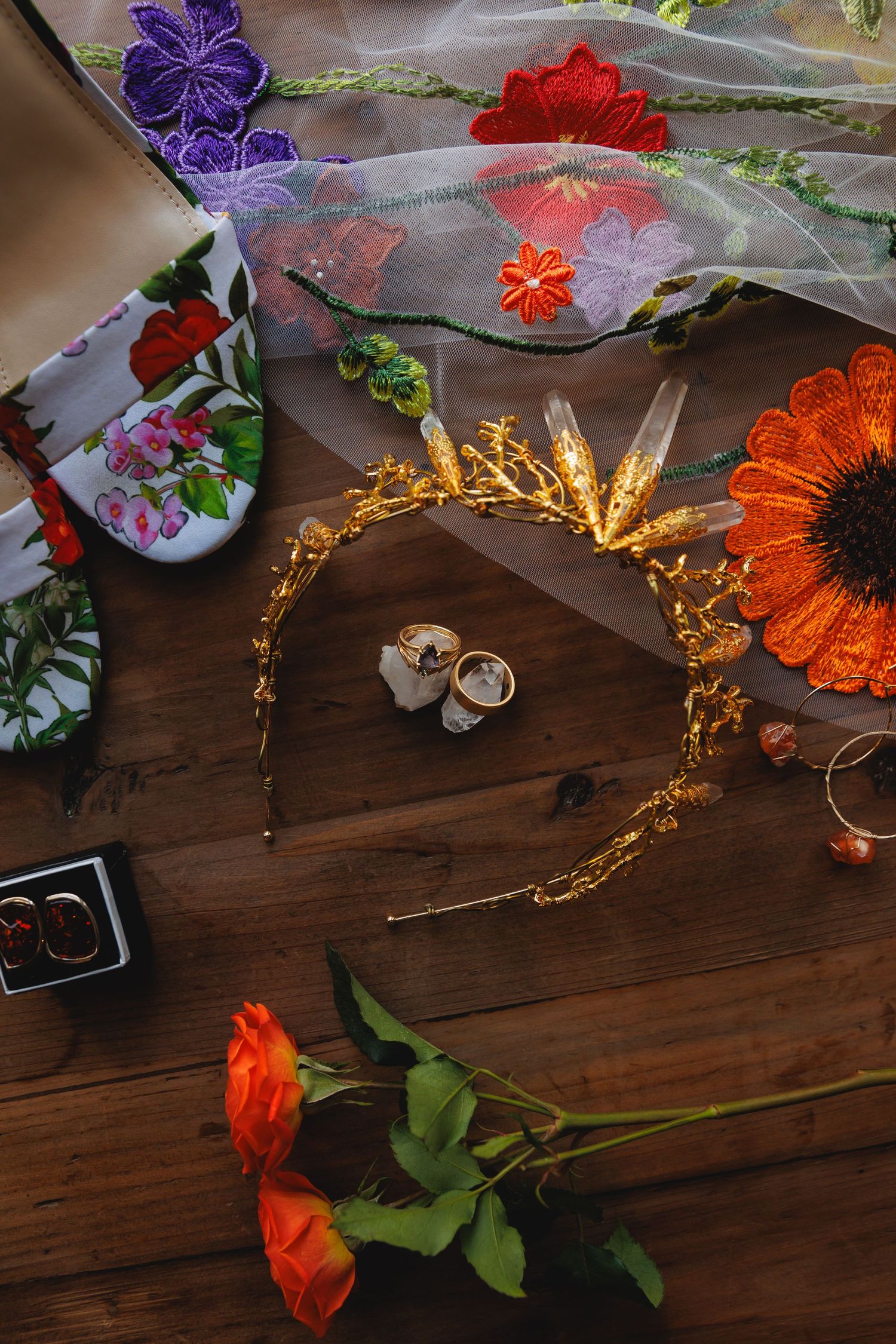 Wedding details with colorful flowers, gold crown, and rings on wooden surface.