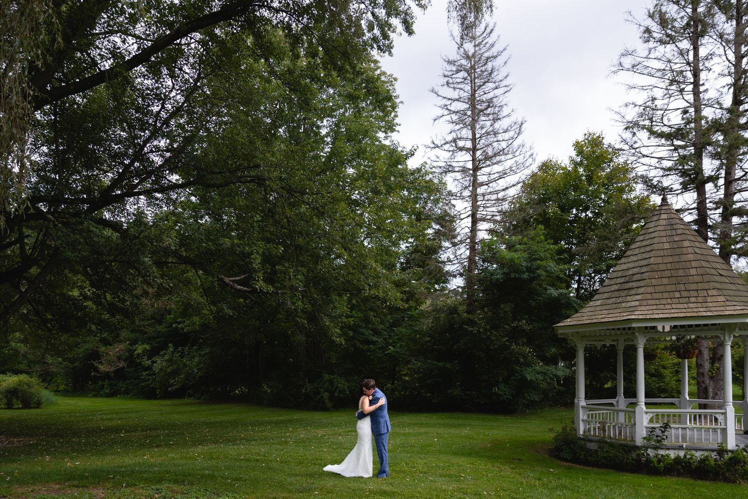 A couple embraces near a white gazebo surrounded by lush green trees and grass in a scenic outdoor setting.