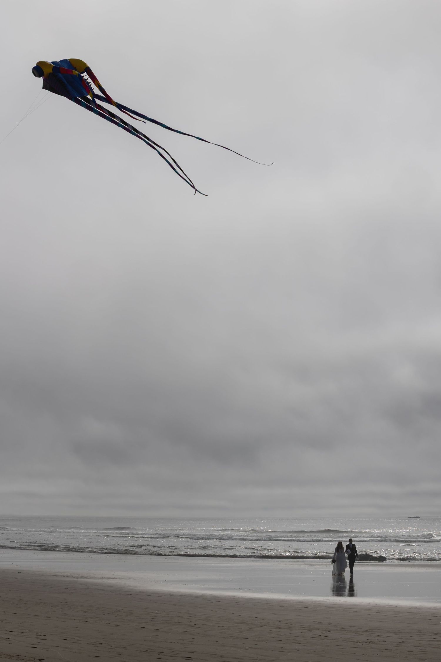 A colorful kite soars above a misty beach where two figures stand by the ocean on a cloudy day.