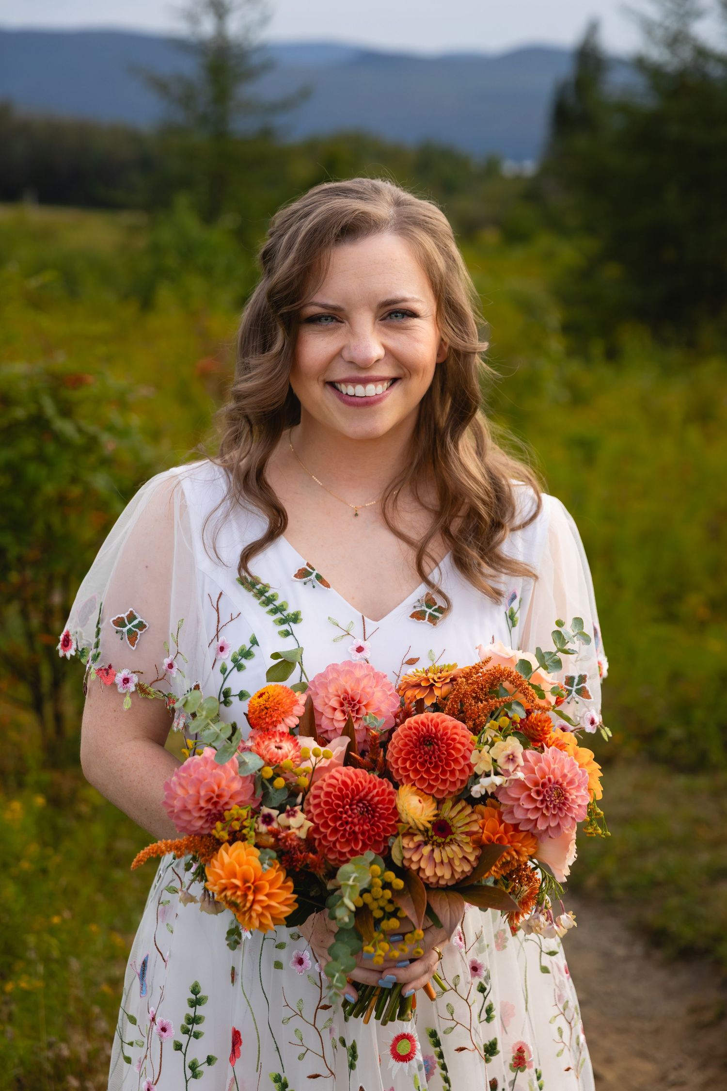 A bright bouquet of orange and coral dahlias complements a delicate white floral dress against a mountain backdrop.