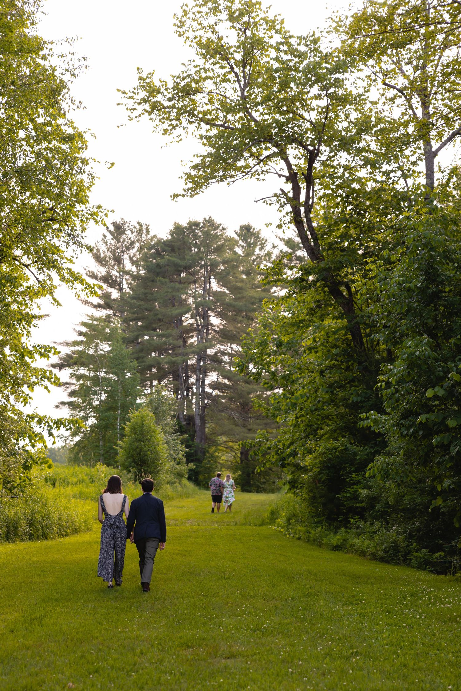 Two people walk along a peaceful grassy path bordered by lush green trees on a summer afternoon.