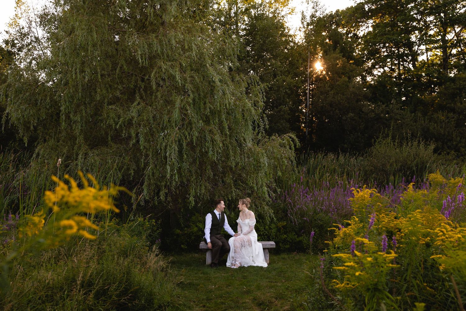 A couple in white attire poses in a garden at sunset with wildflowers and willow trees in the background.
