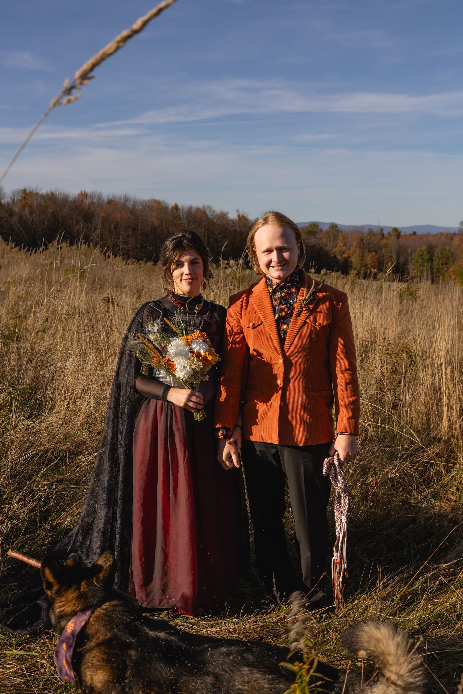 Two people stand together in a fall field with tall grass and autumn trees in the background.