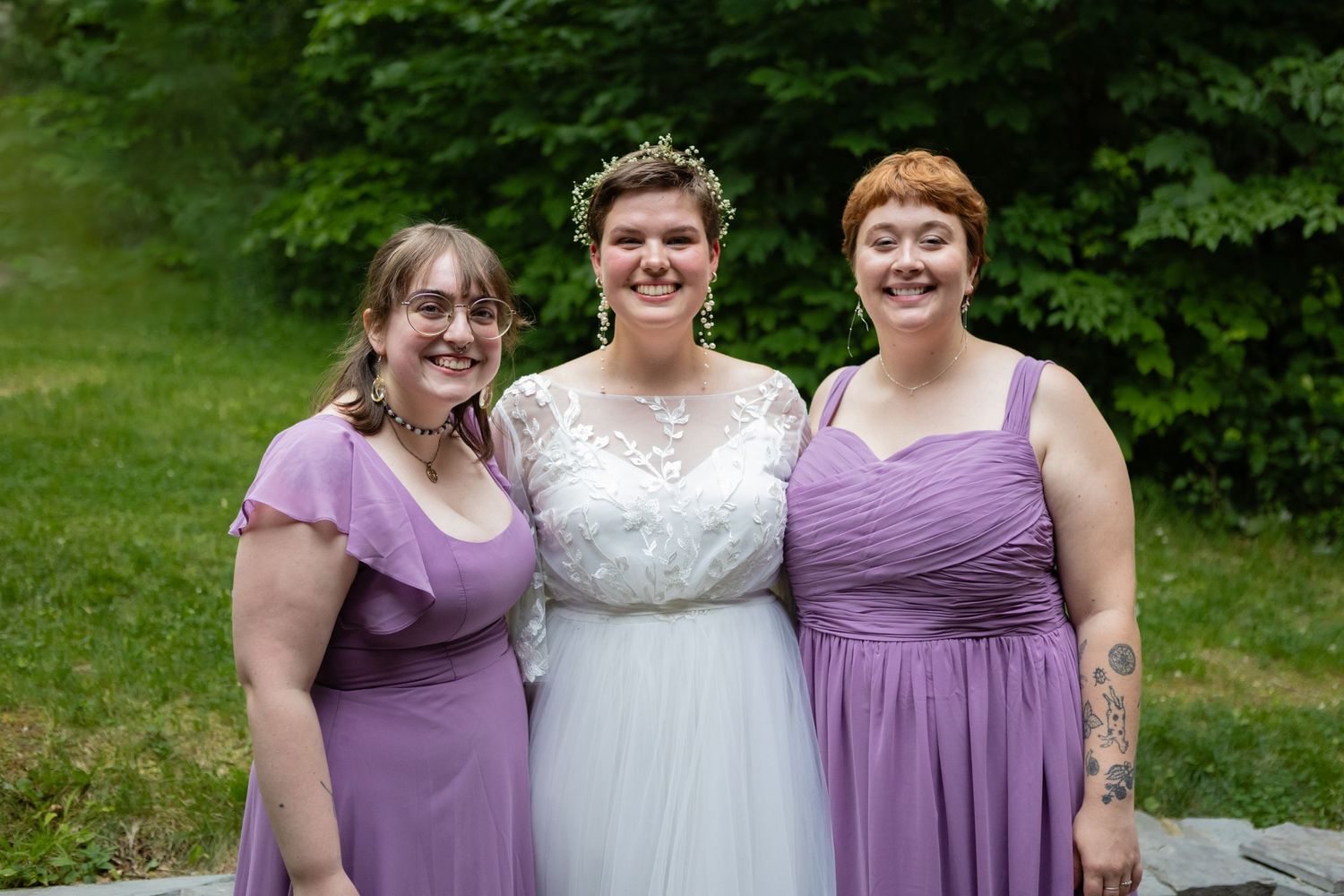 Wedding party in white and purple dresses pose together outdoors against green background.