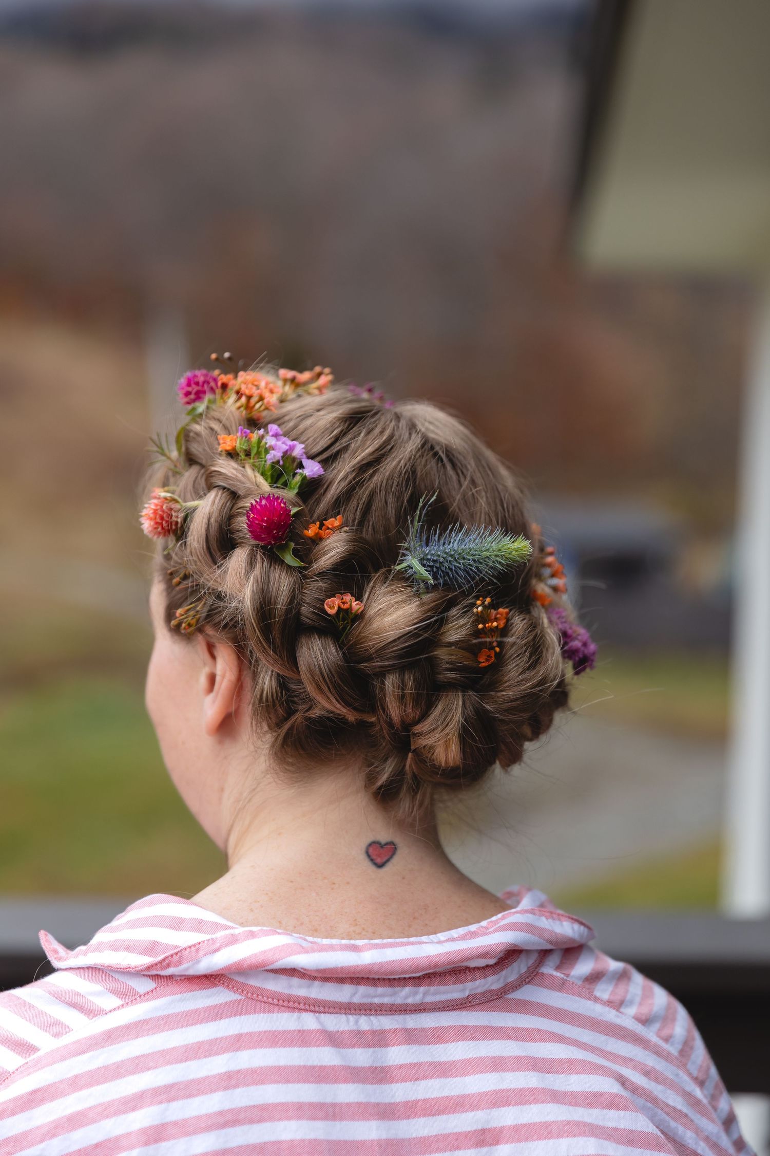 Braided hairstyle adorned with colorful flowers and small heart tattoo visible on neck.