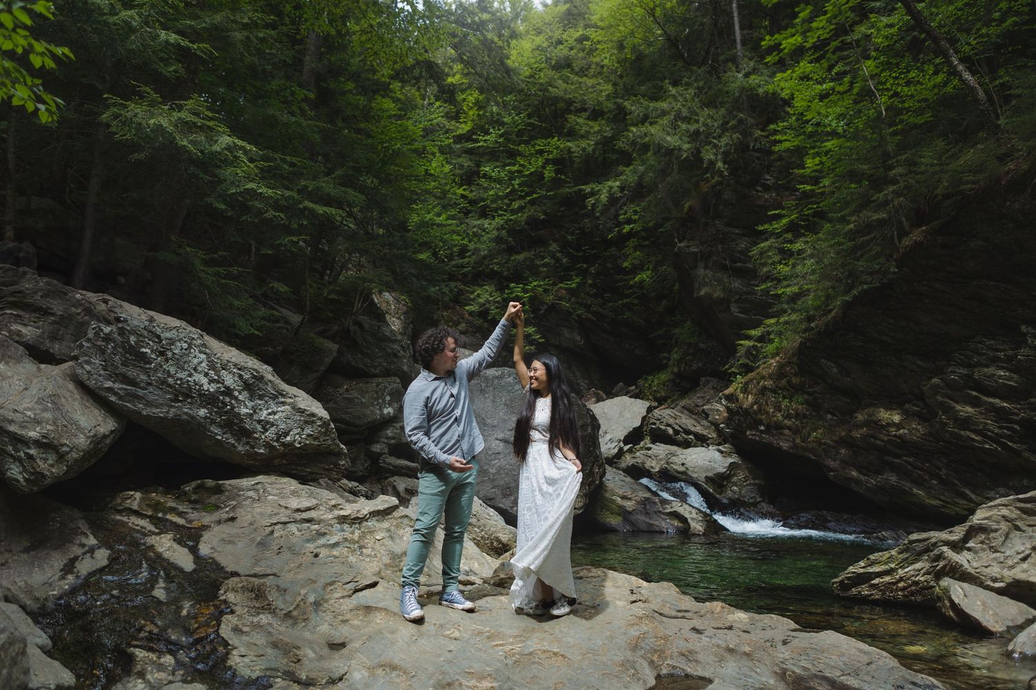 Two people celebrate on rocky terrain near a waterfall in a lush forest setting.