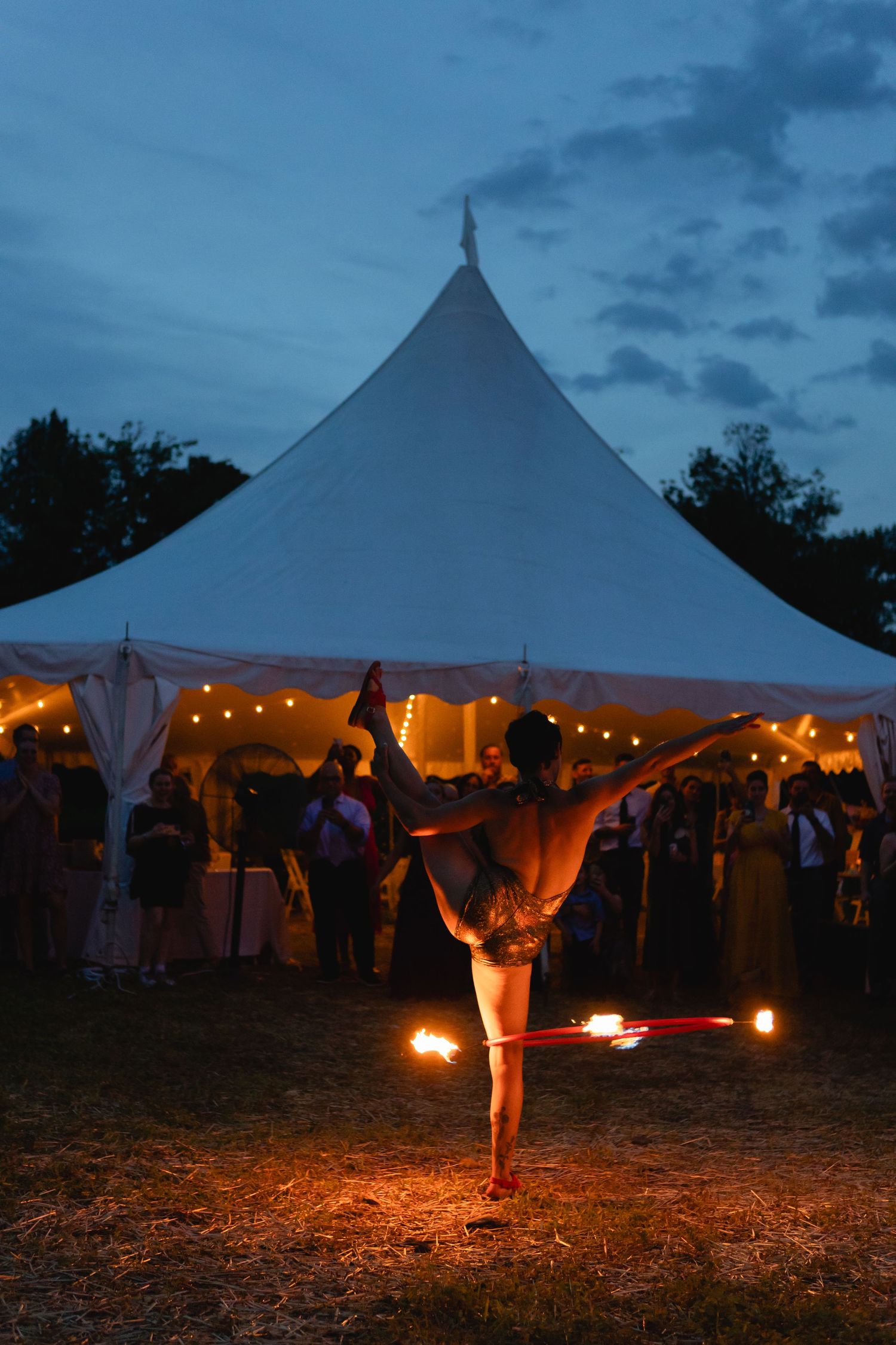 Fire dancer performs at twilight outdoor wedding reception under white tent with string lights.