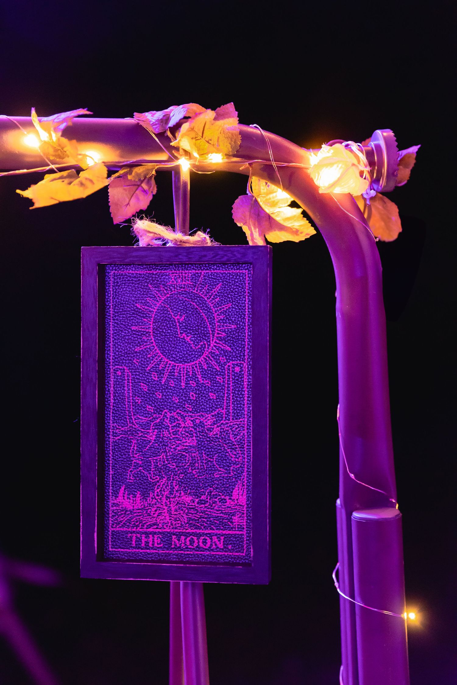 The Moon tarot card displayed in purple neon light with fairy lights and leaves.