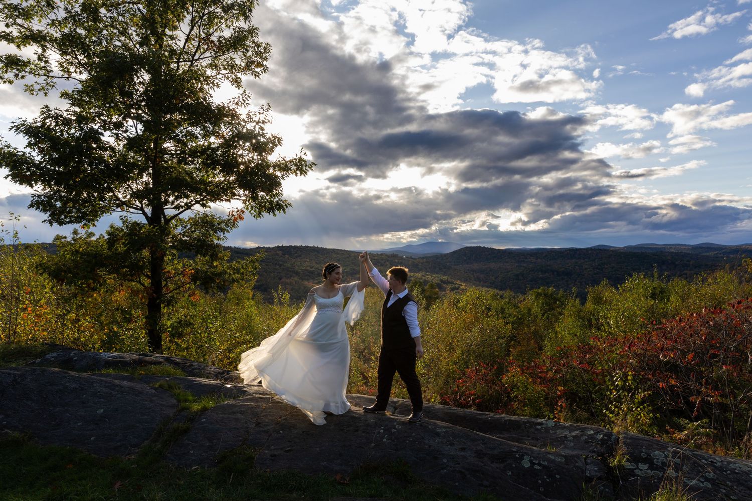 A wedding couple dances on a scenic overlook with dramatic mountain views and clouds at sunset.