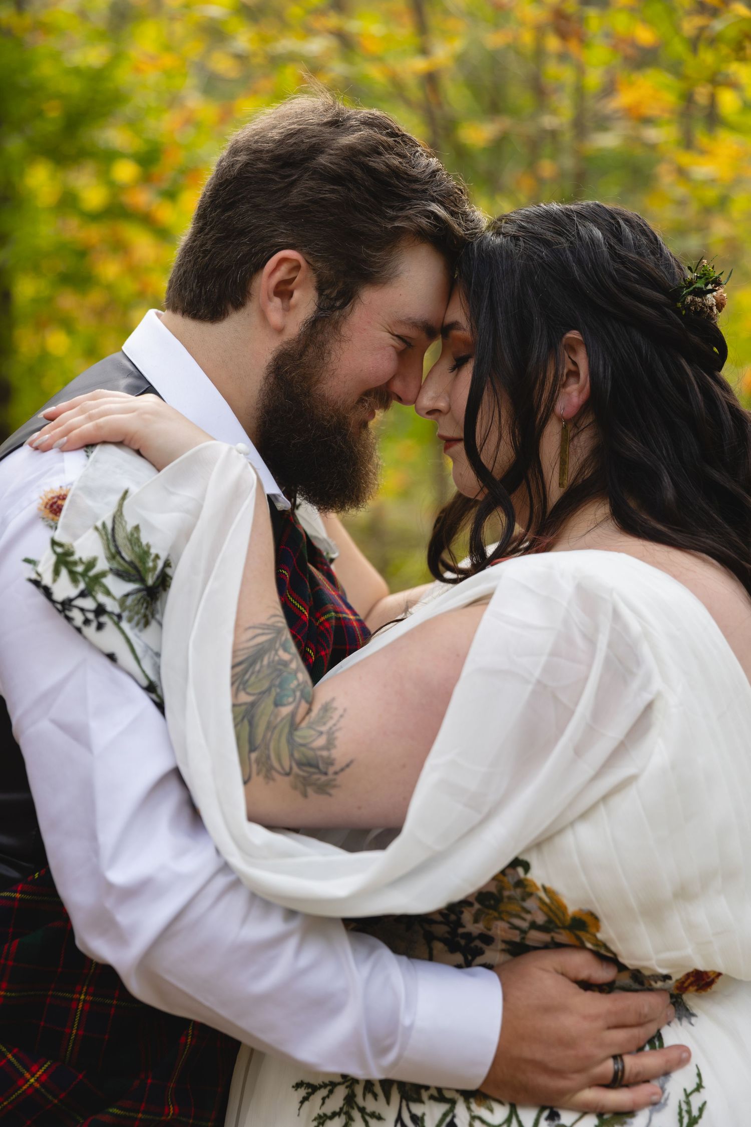 Newlyweds embrace in romantic forest setting with autumn colors in background.