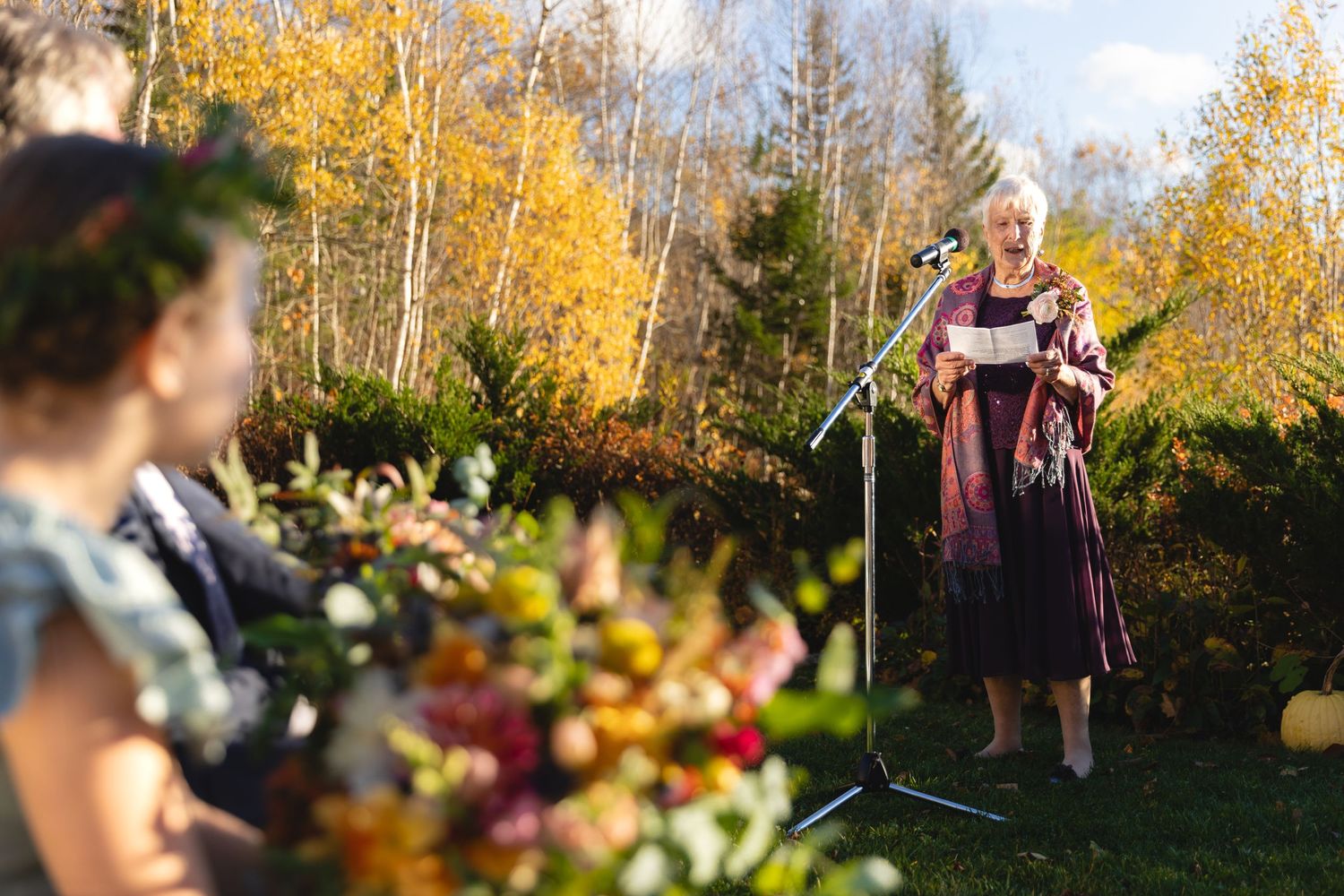 A wedding ceremony takes place outdoors with guests seated amidst colorful fall foliage.