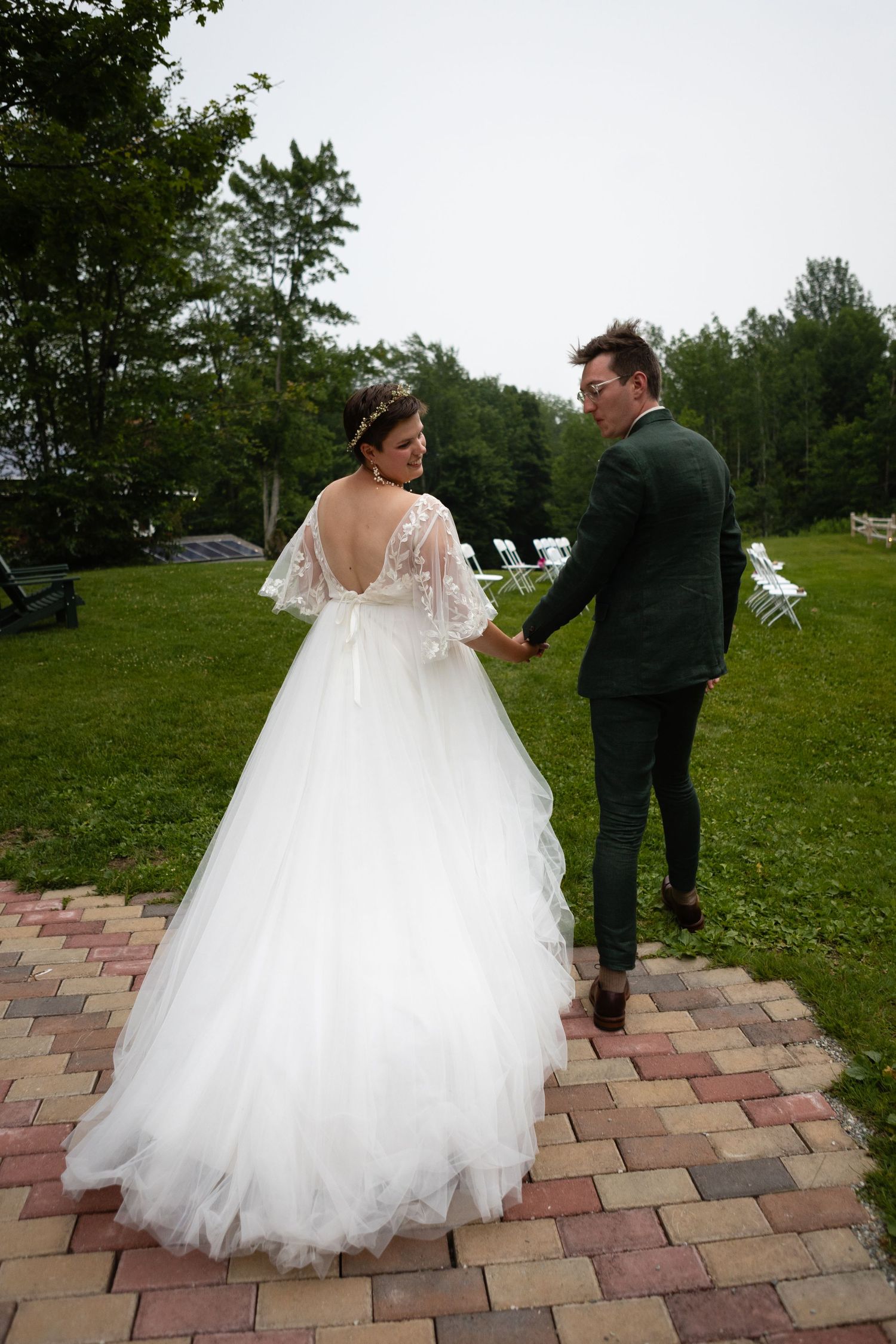 Couple in wedding attire walking on brick pathway with green landscape in background.