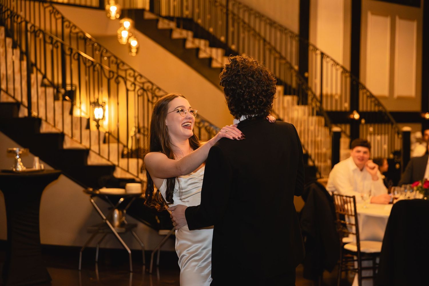 A couple sharing a joyful dance moment at an elegant venue with warm lighting and iron stair railings in the background.