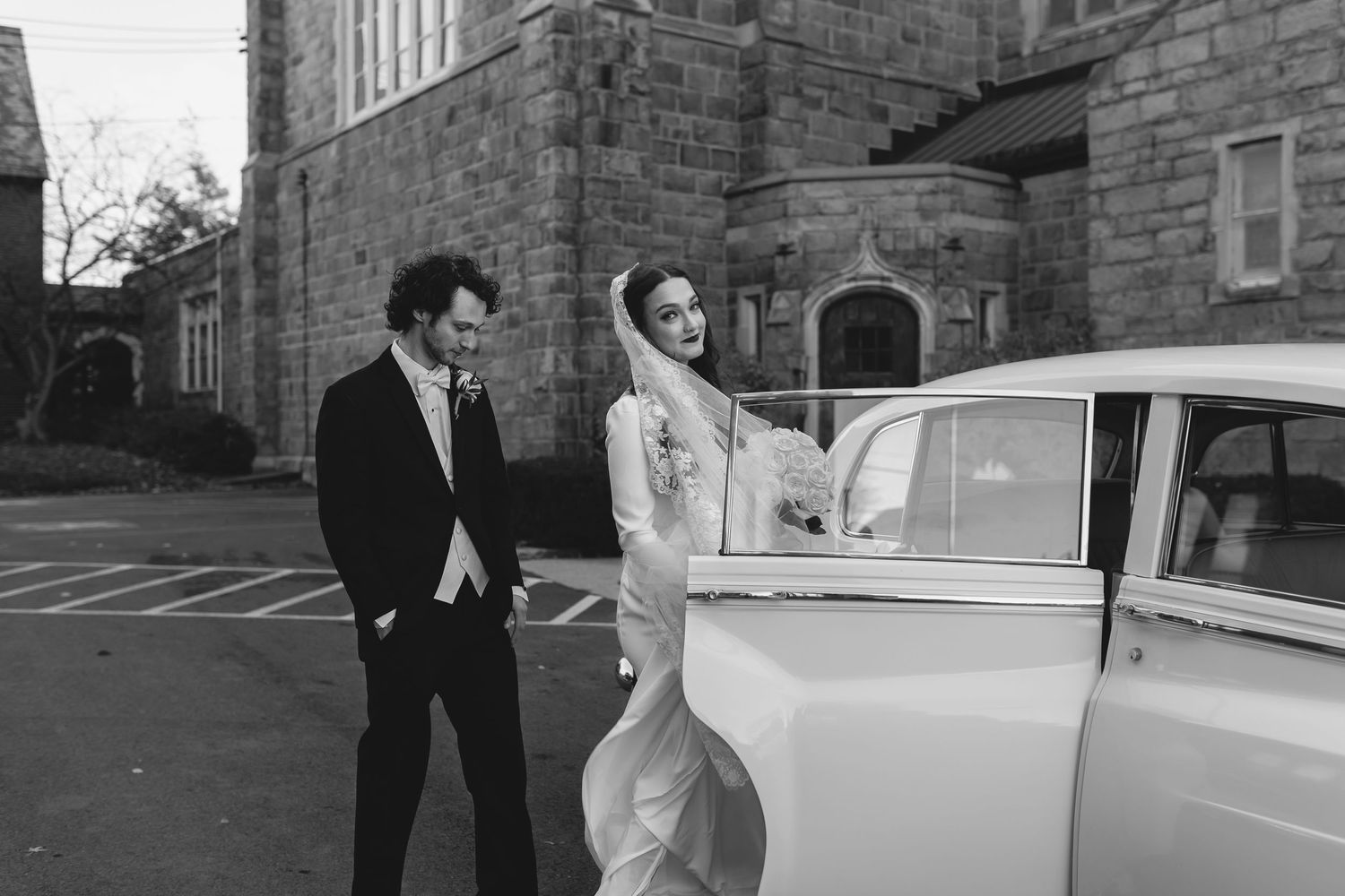 A couple in formal wedding attire stands next to a vintage white car outside a stone building.