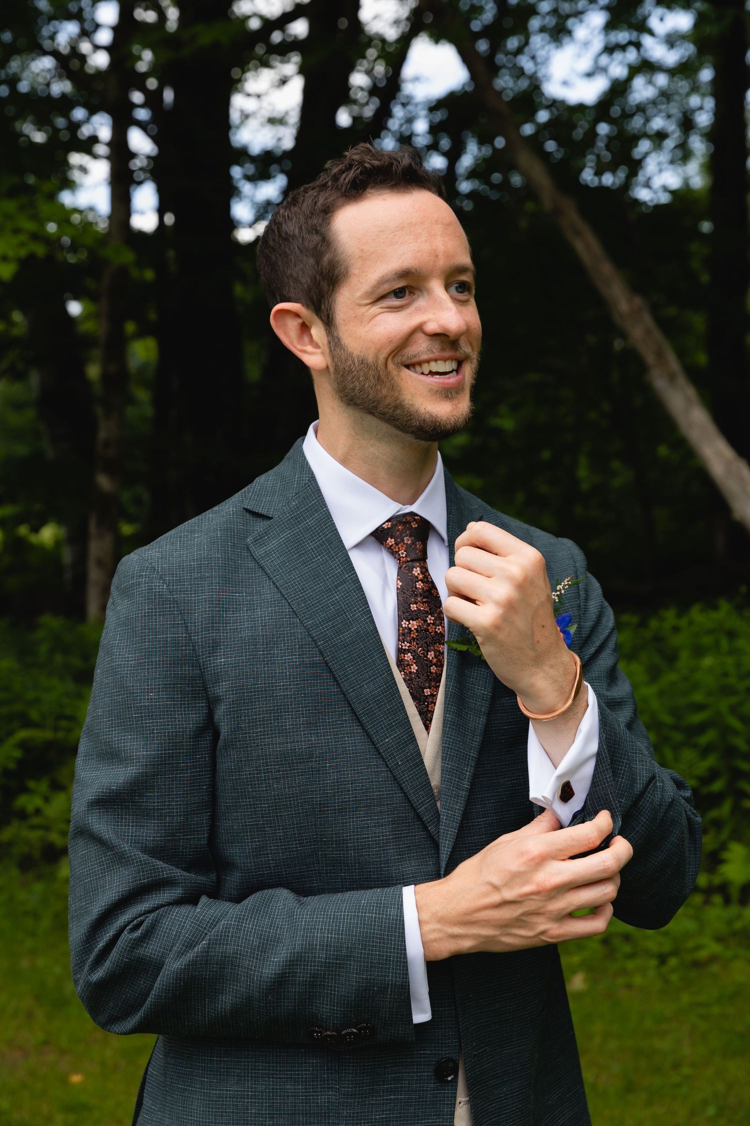 Groom in charcoal suit smiling while adjusting tie outdoors.