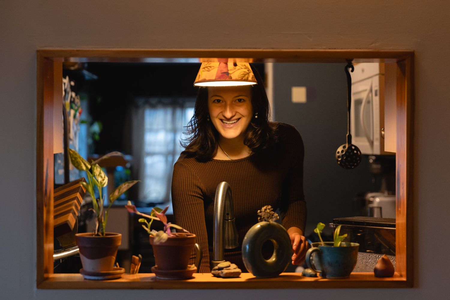 A person in a black sweater stands behind potted houseplants near a window in a dim, cozy interior setting.