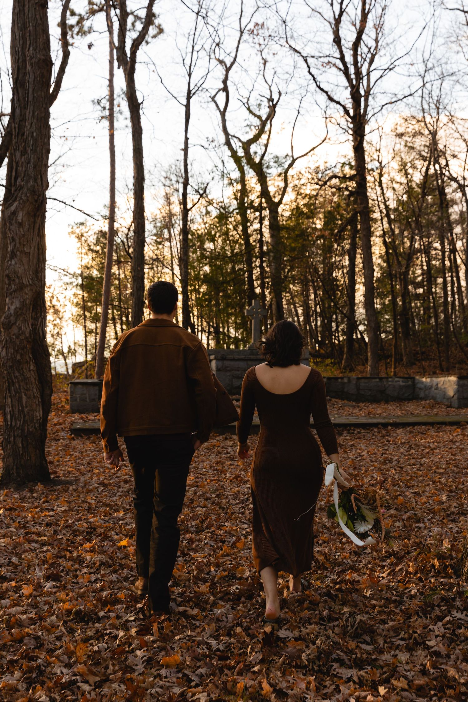 A couple walking through a fall forest with golden leaves scattered on the ground during sunset.