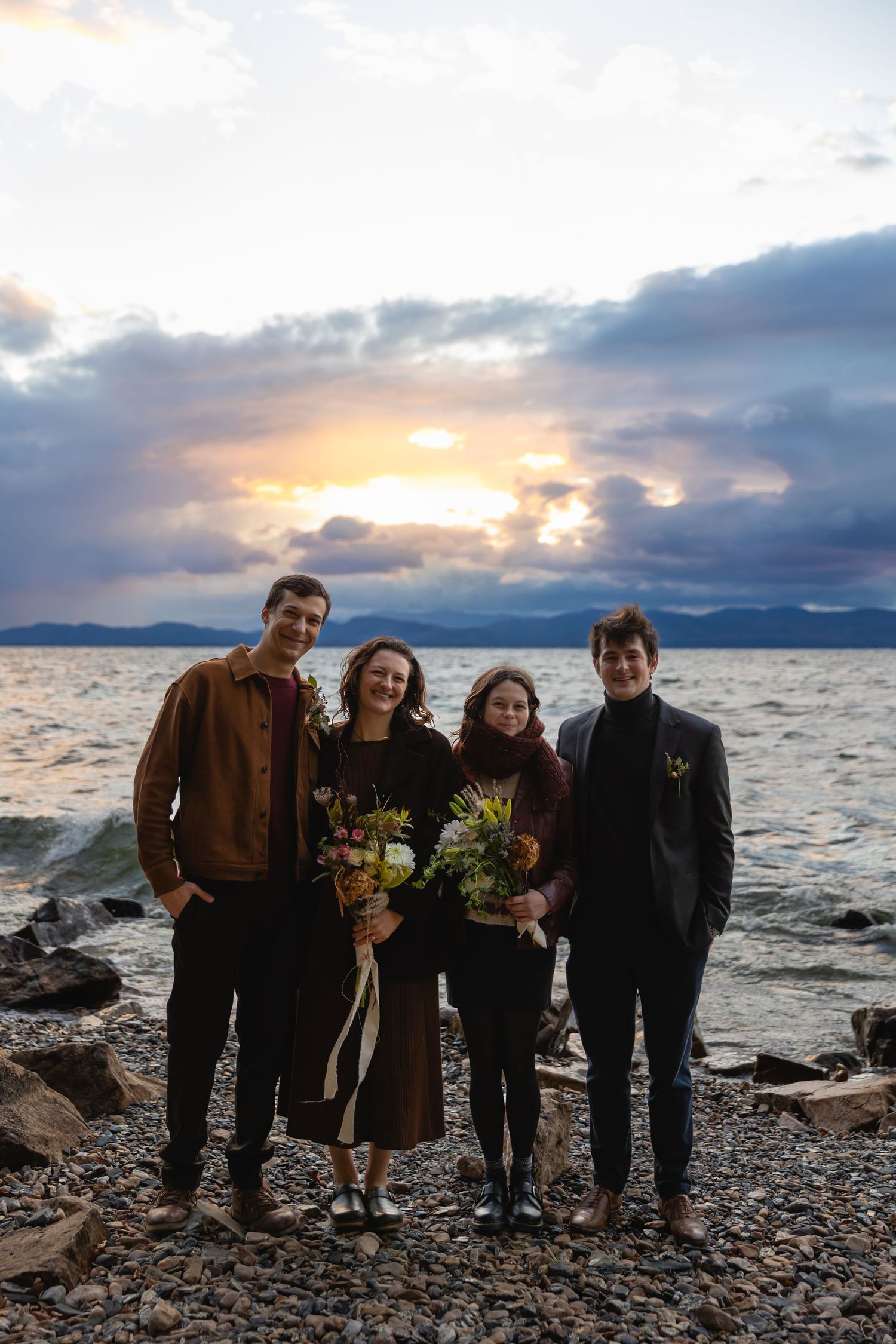 A group of four friends stands together on a rocky beach at sunset holding flower bouquets with mountains in the distance.