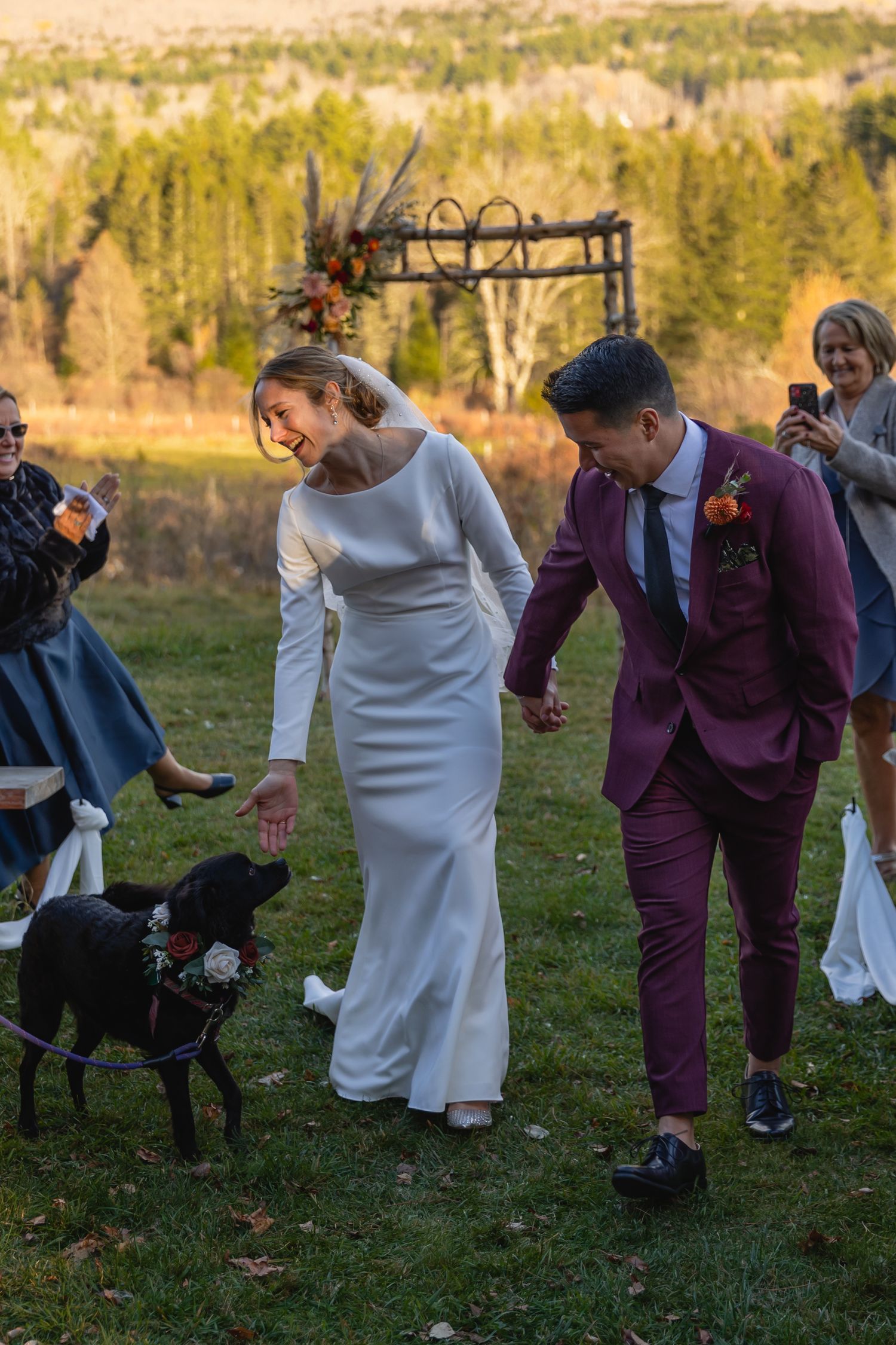 A couple in wedding attire interacts with a black dog during an outdoor autumn ceremony with wooden arch backdrop.