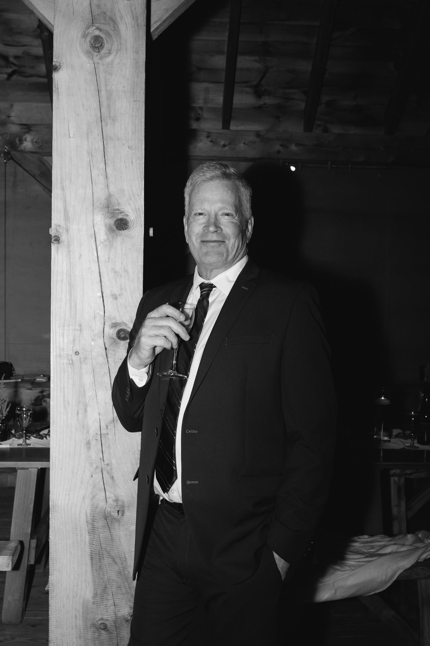 Black and white photo of a person in a formal suit and tie posing against a wooden post in a dimly lit venue.